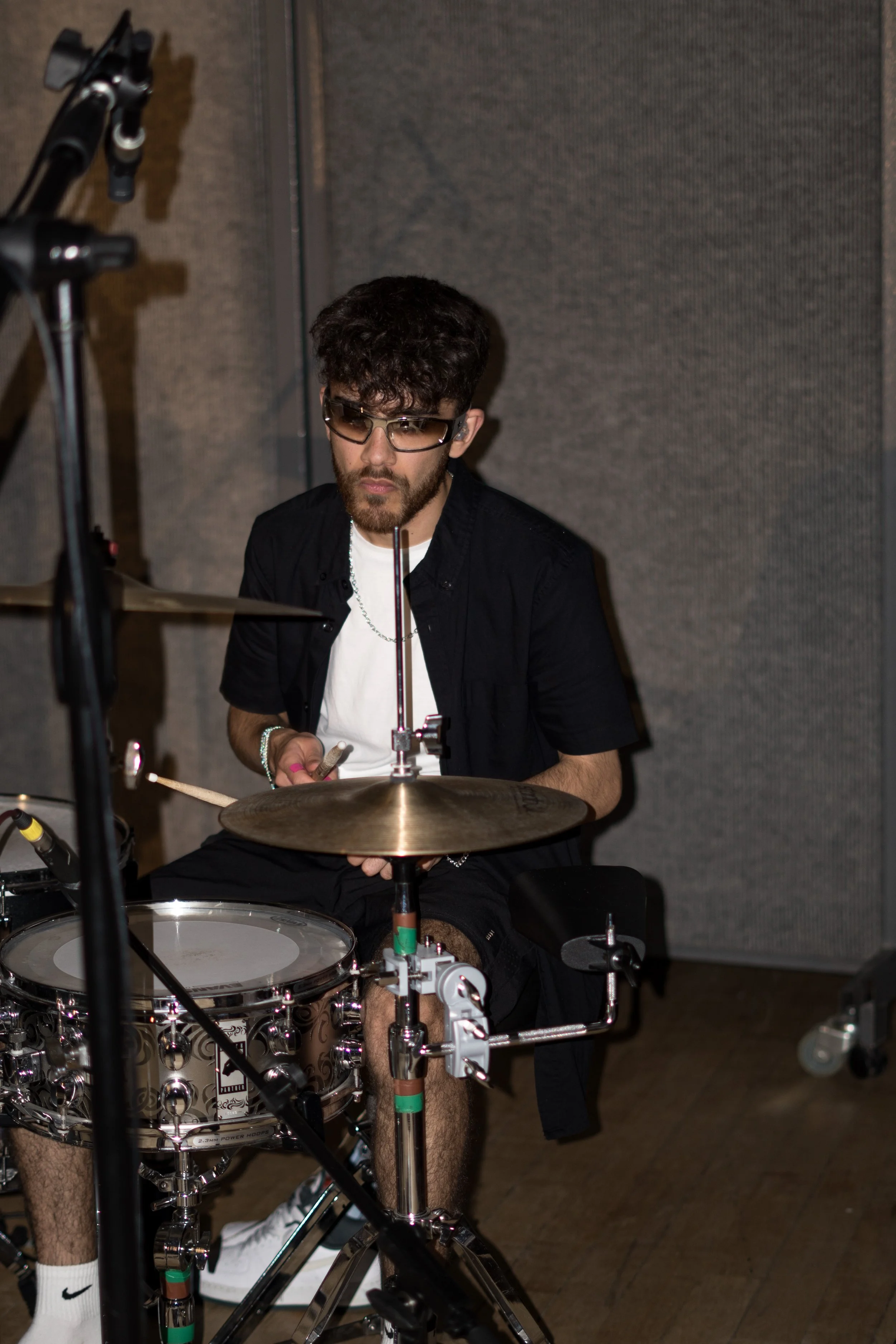 Young man with curly hair and beard playing drums in a recording studio, wearing sunglasses, a black shirt, and white Nike sneakers.