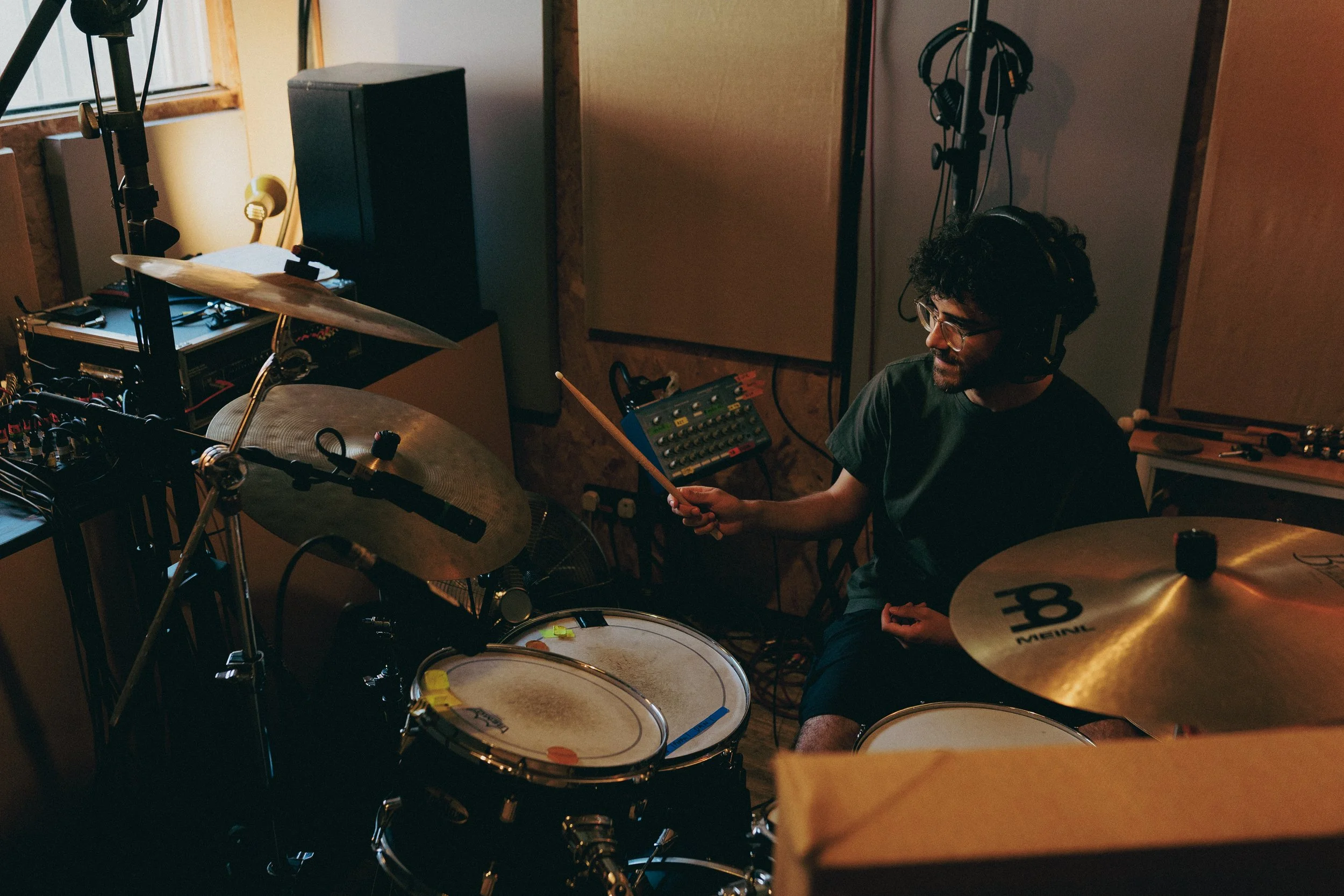A man with curly hair and glasses sitting in a recording studio, surrounded by musical equipment including drums, a speaker, headphones, and recording gear.