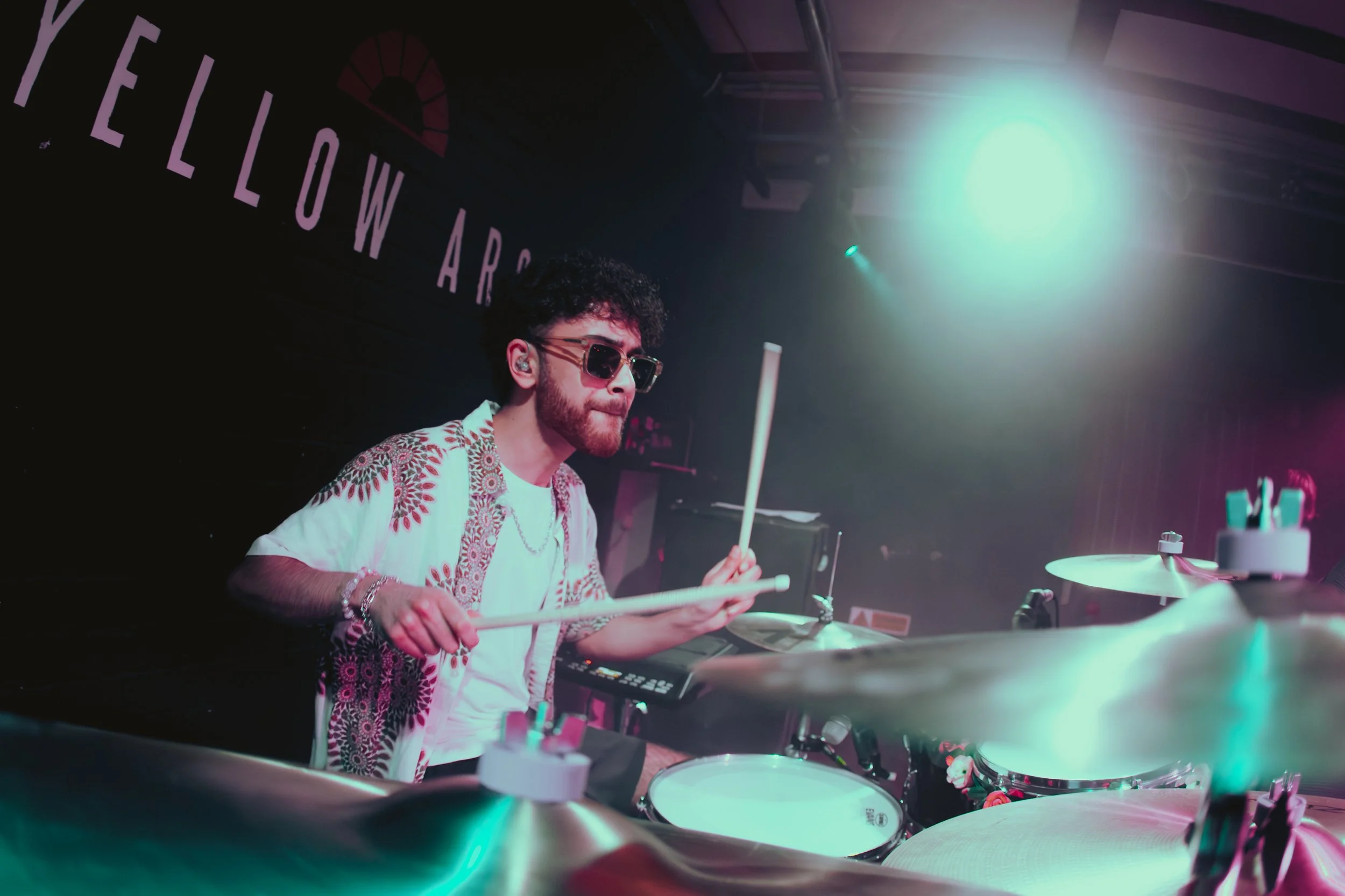 A man playing drums in a dimly lit room with colorful lighting, wearing sunglasses and a patterned shirt.