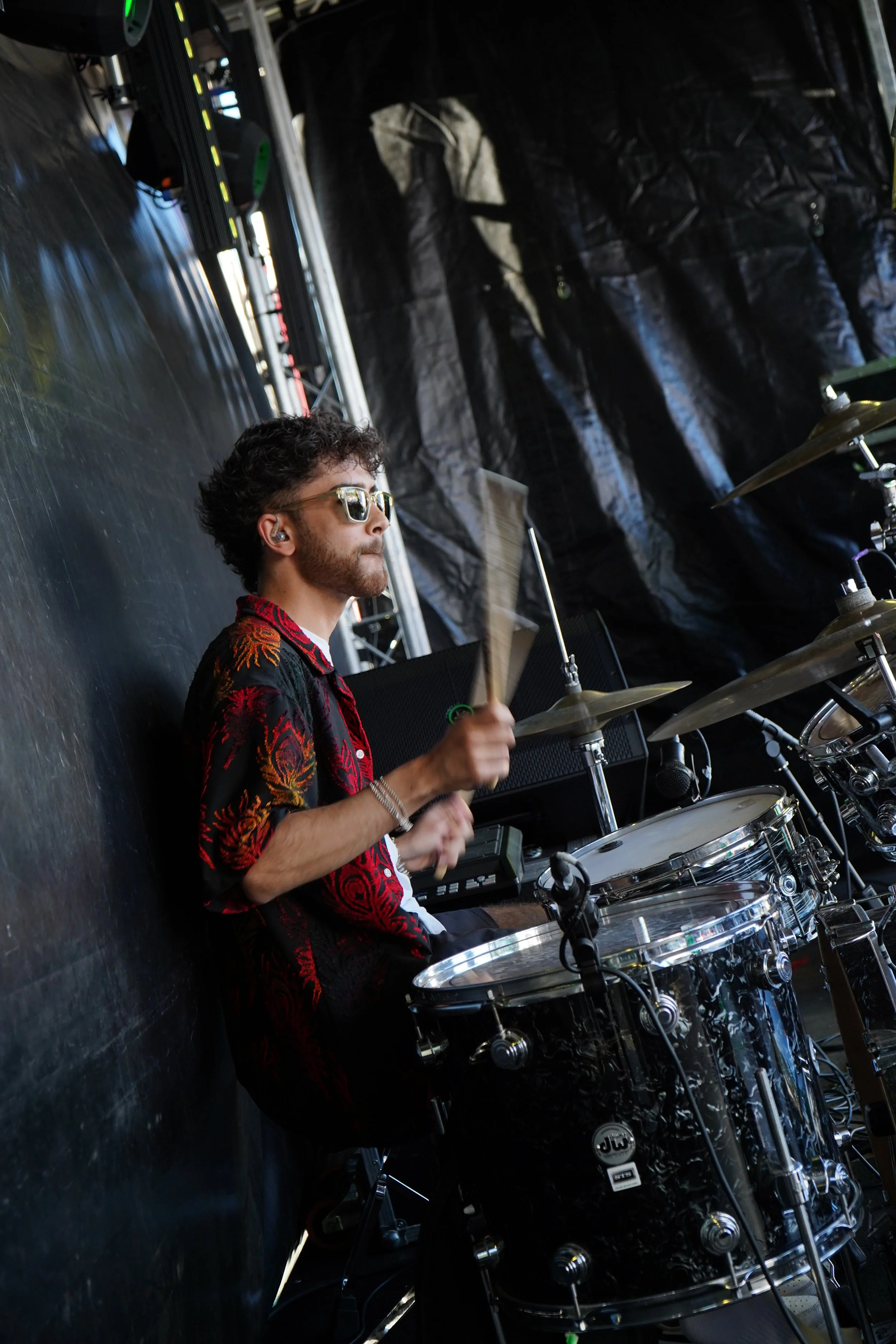 Musician playing drums on stage, wearing sunglasses and a black and red shirt, with a drum set and stage equipment around him.