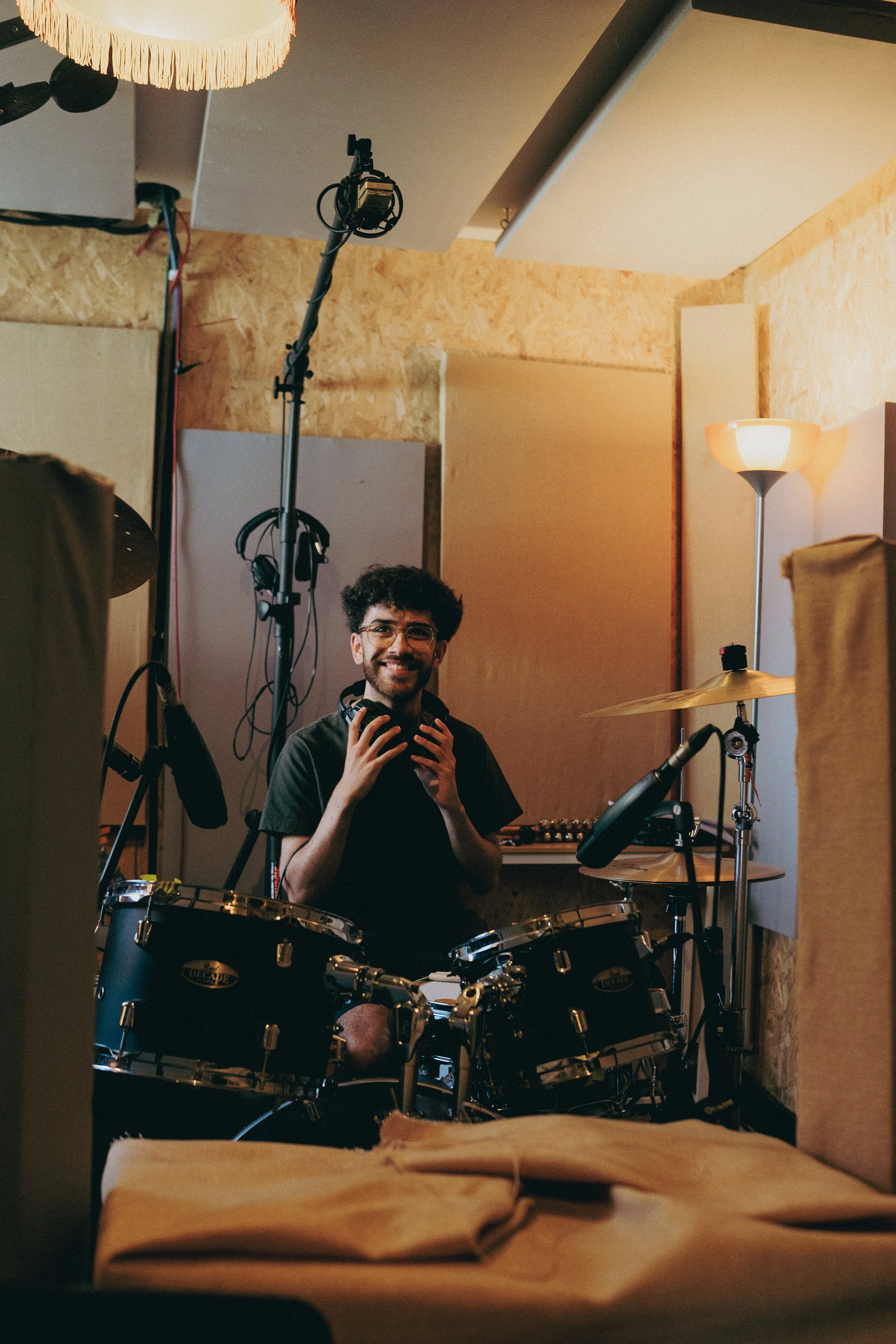 A young man with glasses and a beard is sitting behind a drum set in a recording studio or rehearsal space, smiling and holding headphones. The room has soundproofing panels, a floor lamp, and music equipment.