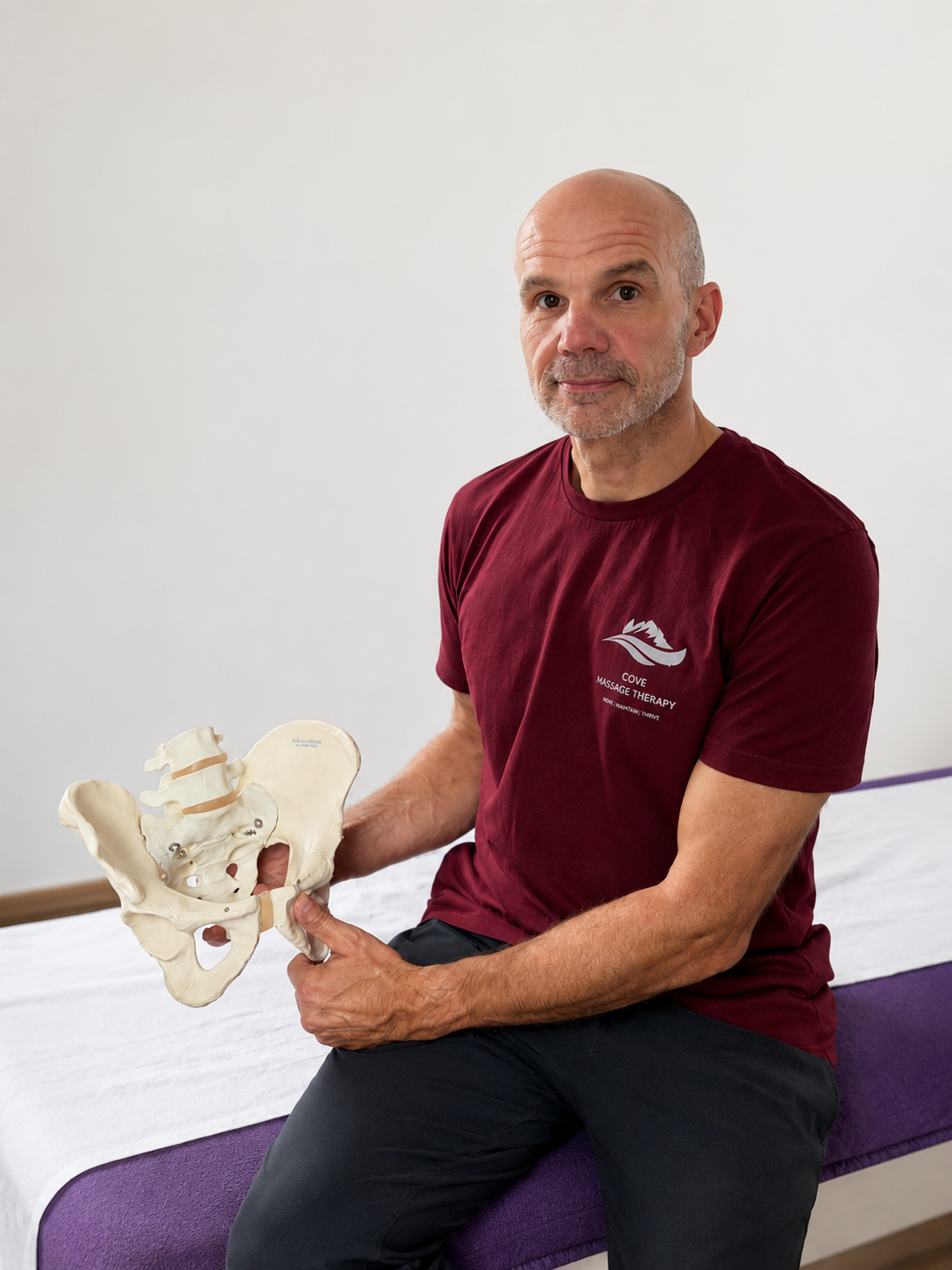 A man wearing a maroon shirt with 'Cove Massage Therapy' logo sits on a purple examination table holding a medical model of a human pelvis in his right hand.