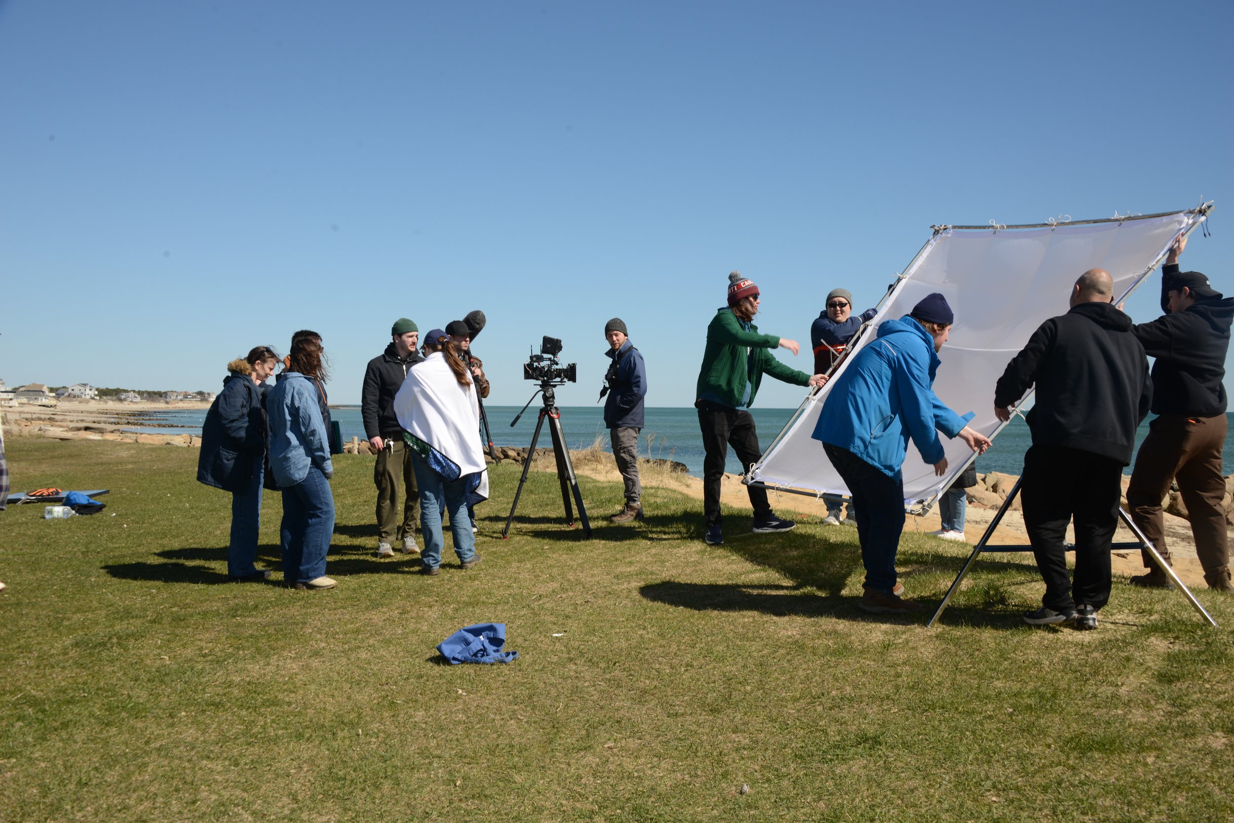 A group of people working together outdoors on a film or photo shoot near the ocean, with some adjusting a large reflector and a camera set up on a tripod, on a grassy area under a clear blue sky.