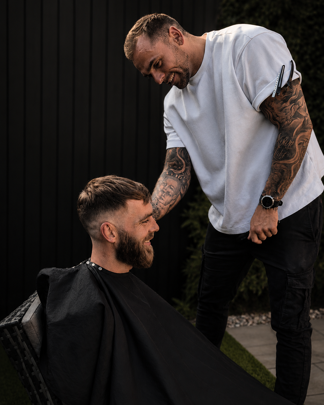 A barber standing and smiling while giving a haircut to a seated man who is also smiling. The barber has tattoos on both arms and is wearing a white T-shirt and black pants. The man getting a haircut has a beard and a short haircut, and a black cape draped over him. They are outdoors against a black fence with greenery.
