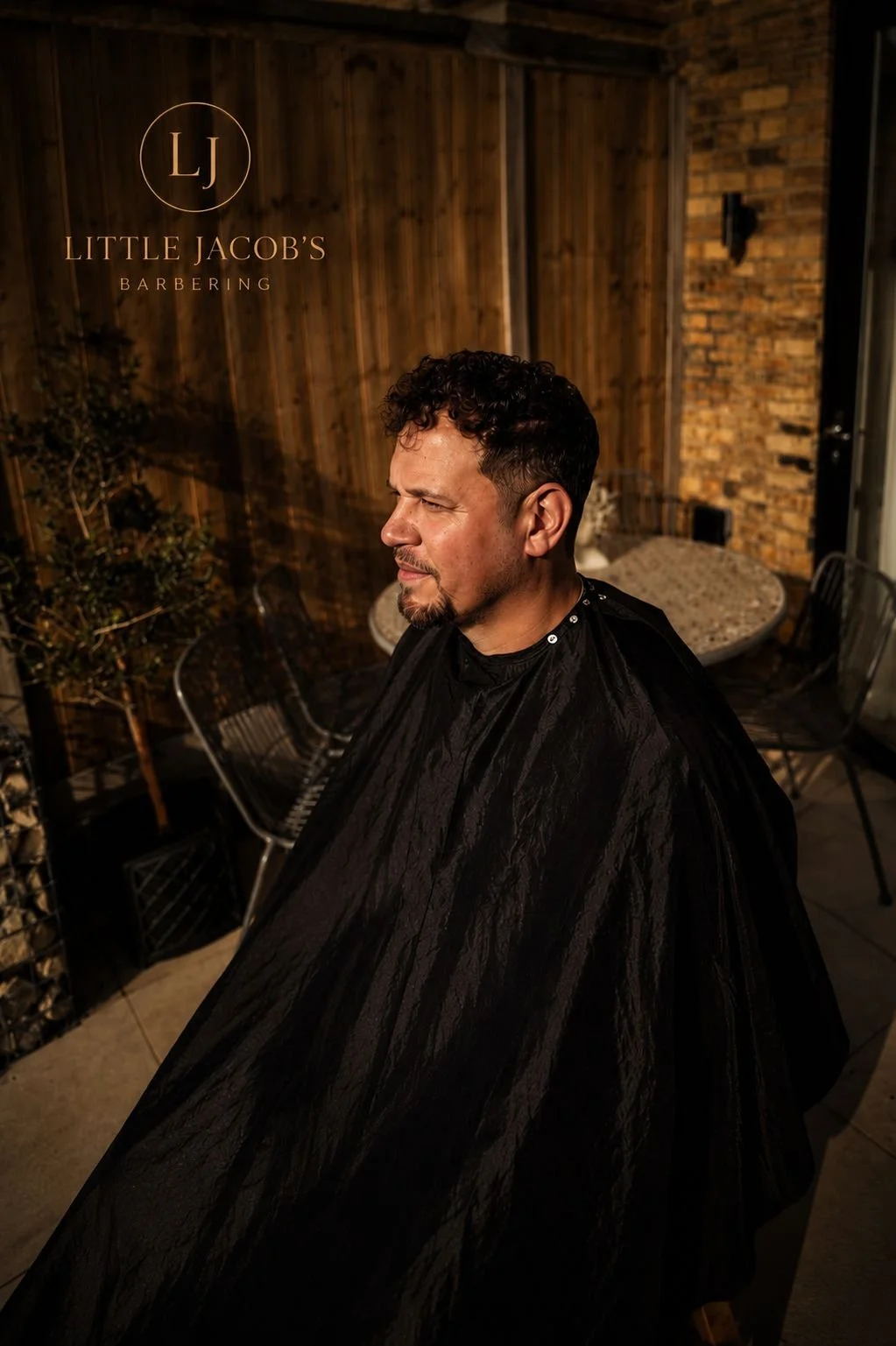 A man with dark curly hair and a goatee sits in a barbershop chair, wearing a black barber cape, at Little Jacob's Barbering, with a wooden and brick wall background.