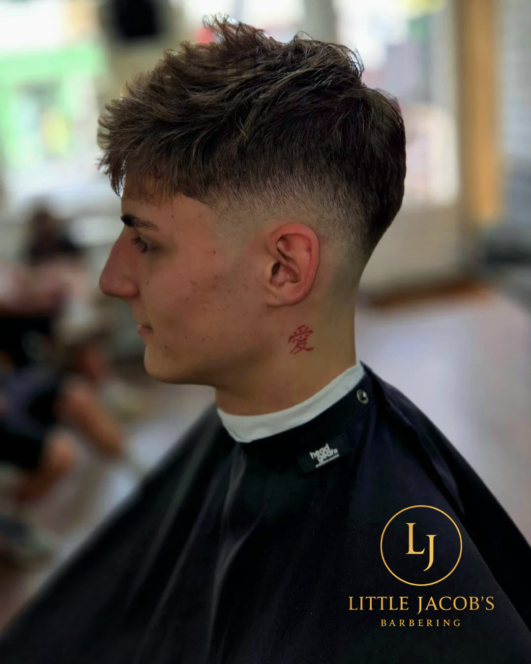 Side profile of a young man with a fresh fade haircut, tattoo on his neck, and wearing a barber cape at Little Jacob's Barbering salon.