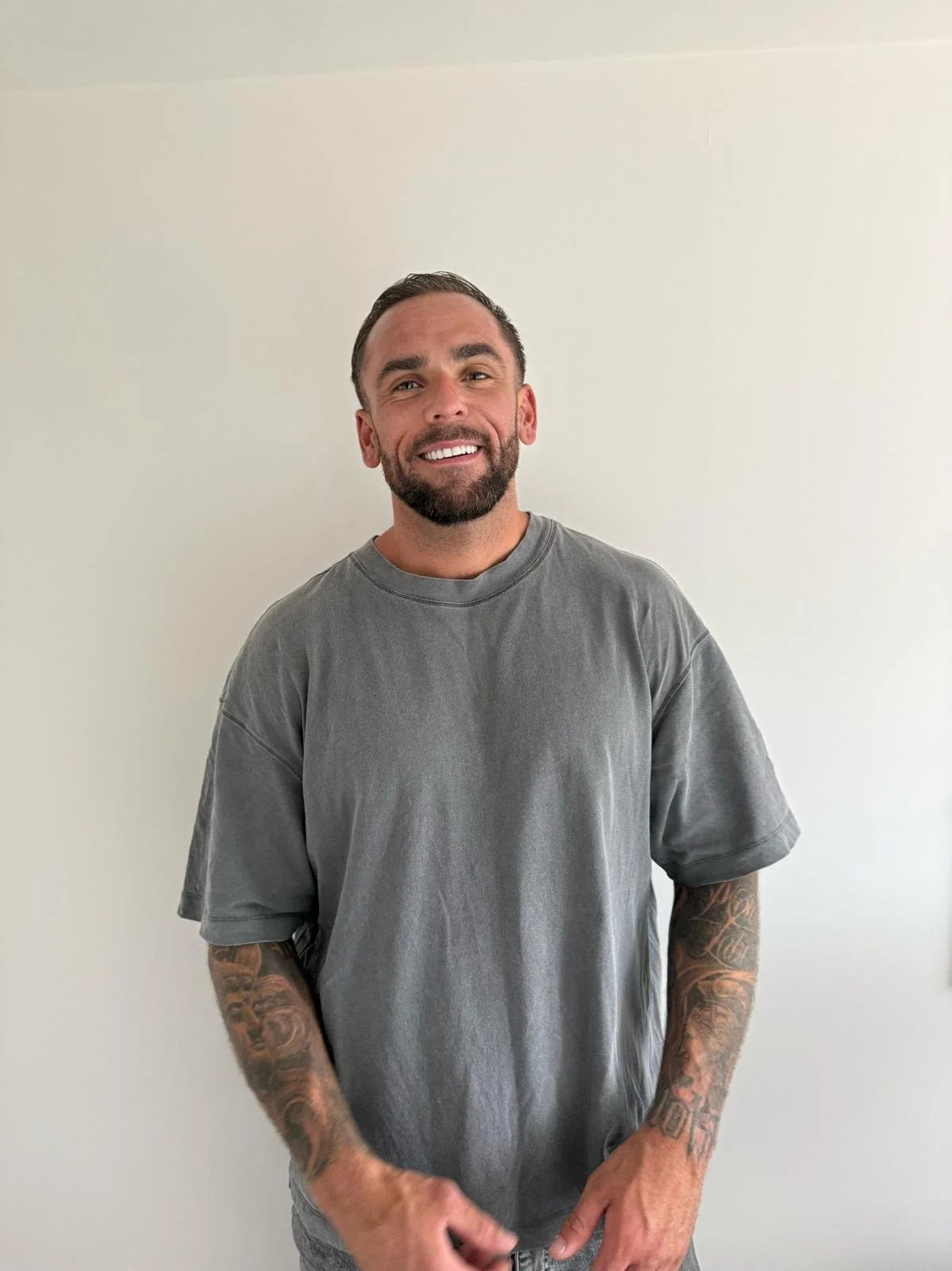 A smiling man with short dark hair and a beard, wearing a loose gray t-shirt, standing against a plain white wall.