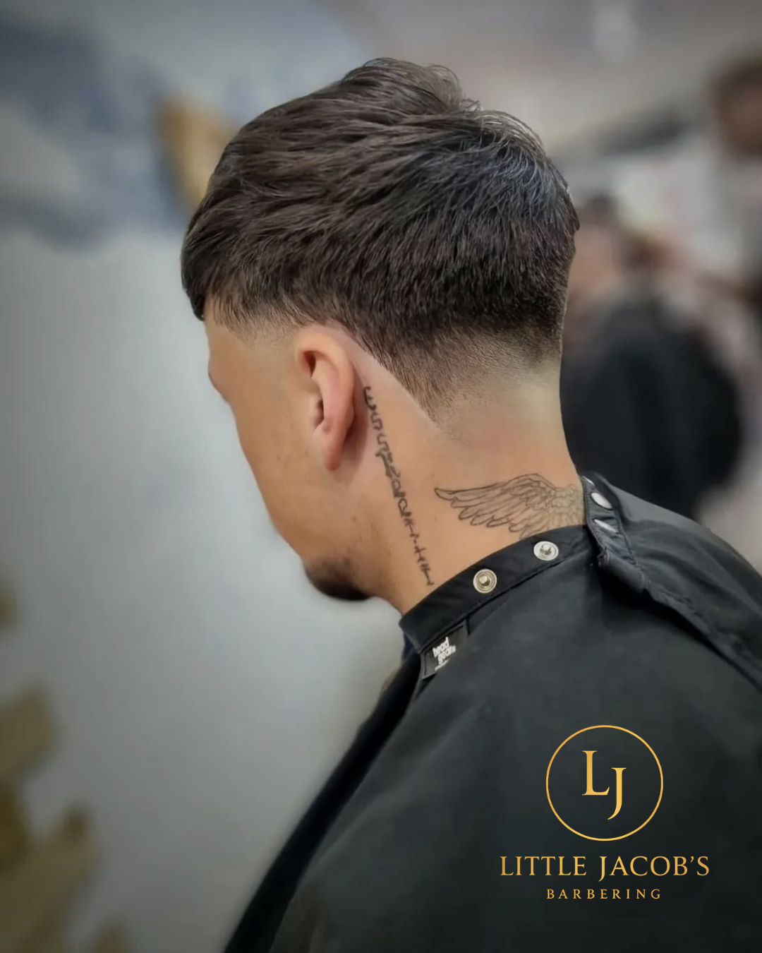 Side view of a man with a modern undercut hairstyle, tattoo behind his ear and on his neck, wearing a barber cape at Little Jacob's Barbering.