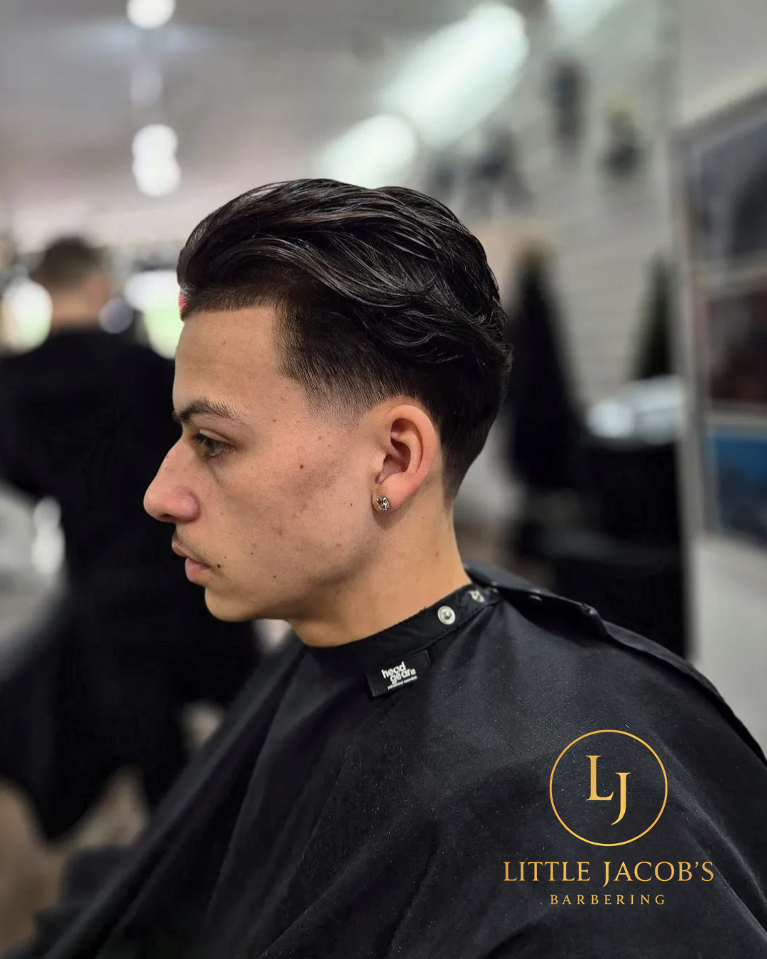 Young man with dark, flyaway styled hair, and clean fade haircut, sitting in a barbershop. He is wearing a black barber cape with a 'LITTLE JACOB'S BARBERING' logo on it.