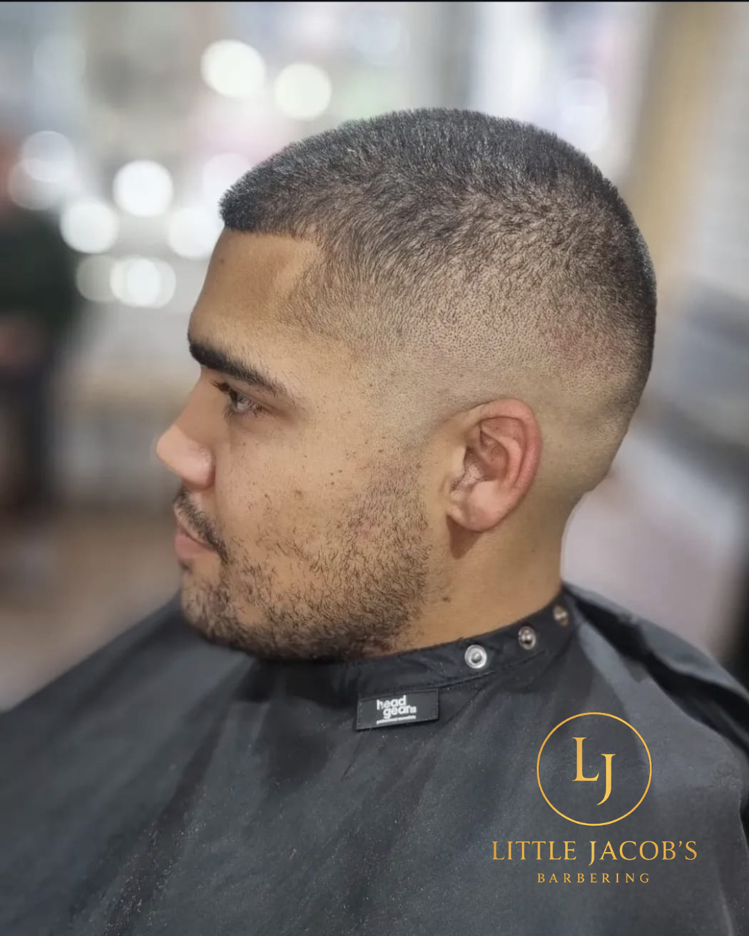 A man with a fresh short fade haircut, facial hair, and wearing a barber cape in a barbershop. The text overlay reads "Little Jacob's Barbering" with a logo.