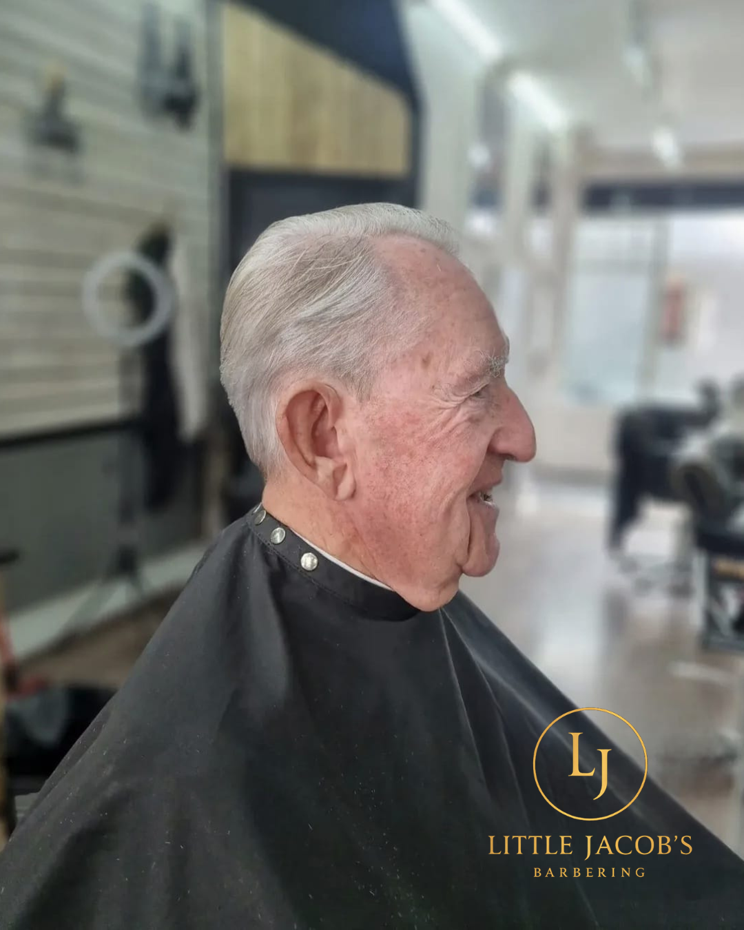 An elderly man with gray hair smiling in a barbershop while getting a haircut, wearing a black barber cape with silver studs, inside Little Jacob's Barbering shop.