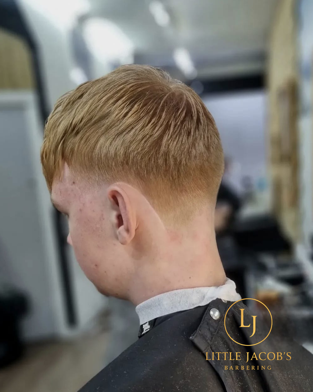 Side view of a young man with short, styled red hair in a barbershop, wearing a black barber cape with the Little Jacob's Barbers logo.