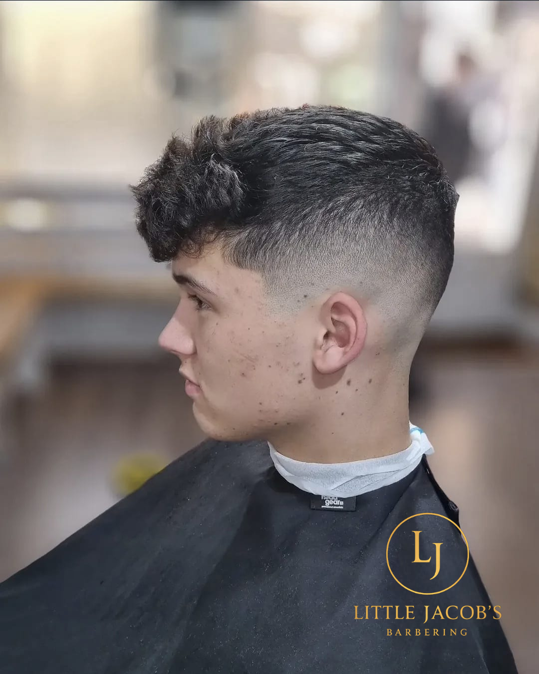 Close-up side view of a young man with a fresh textured fade haircut, short on the sides and longer on top, in a barbershop setting.