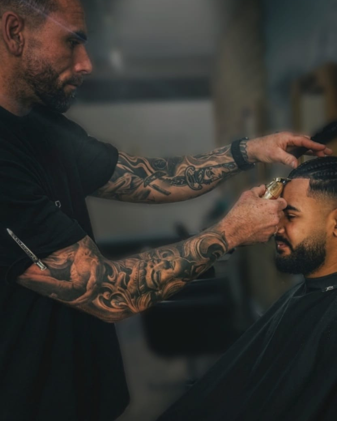 Barber with tattooed arms giving a haircut to a customer with braided hair in a barbershop.