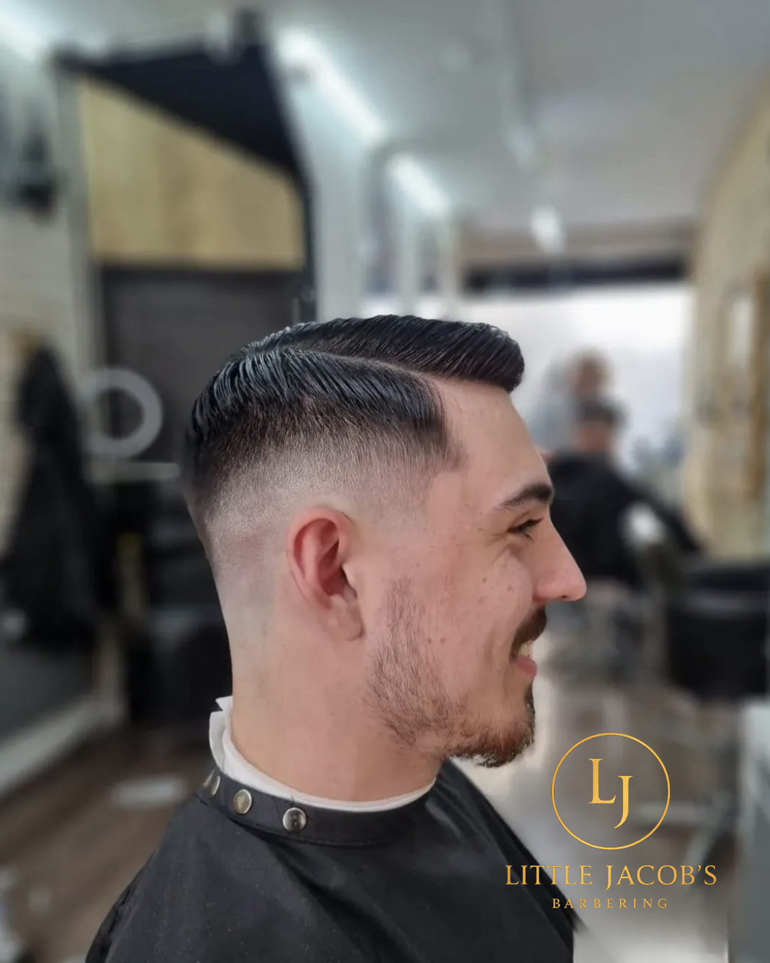 Side view of a young man with a textured fade haircut, styled with a comb-over and a side part, sitting in a barber shop chair, smiling.