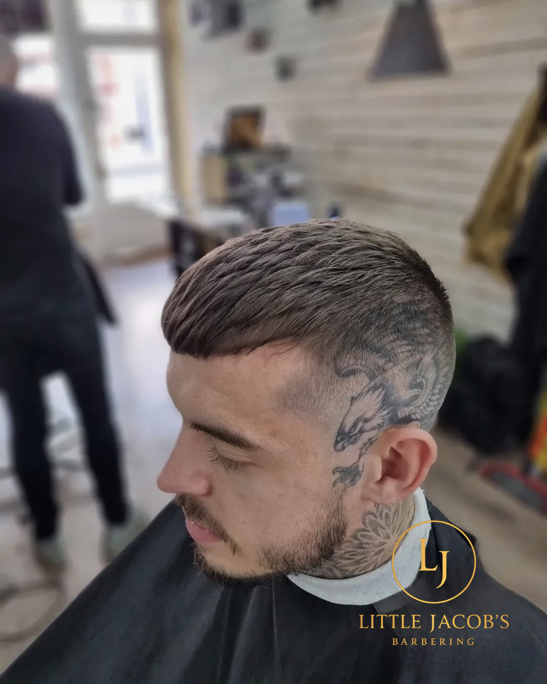 Side profile of a man with a fresh undercut hairstyle, featuring a detailed tattoo of a dragon on the side of his head and neck, inside a barbershop.