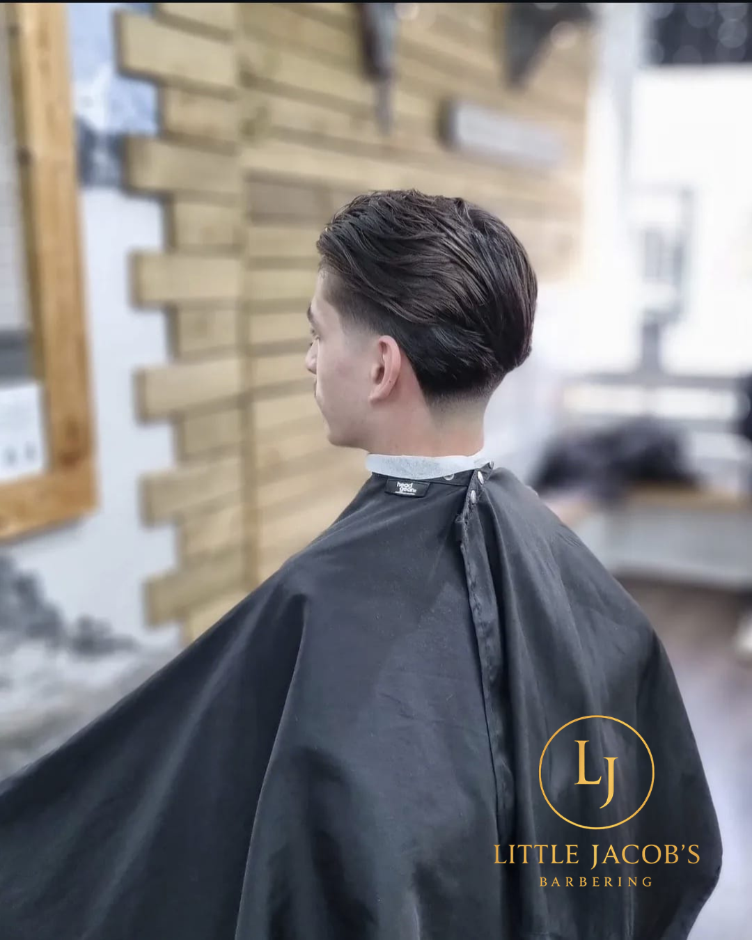 Side view of a man with a stylish, textured haircut in a barber shop, wearing a black barber cape with the logo "Little Jacob's Barbering".