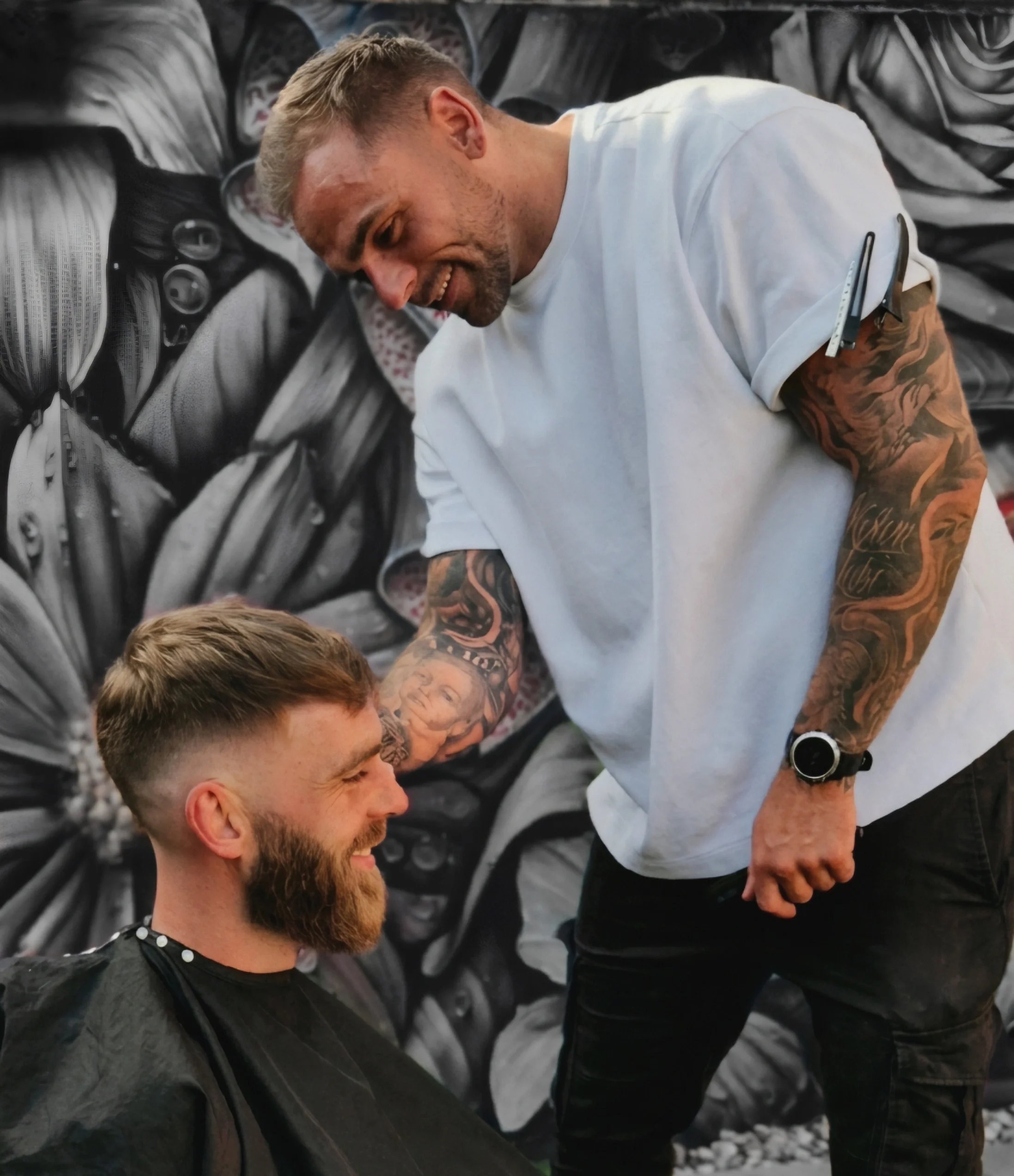 A man with a beard and tattoos on his arms is smiling and facing a young man with short hair and a beard, in what appears to be a barbershop or hair salon. The background features black and white artistic wall art.