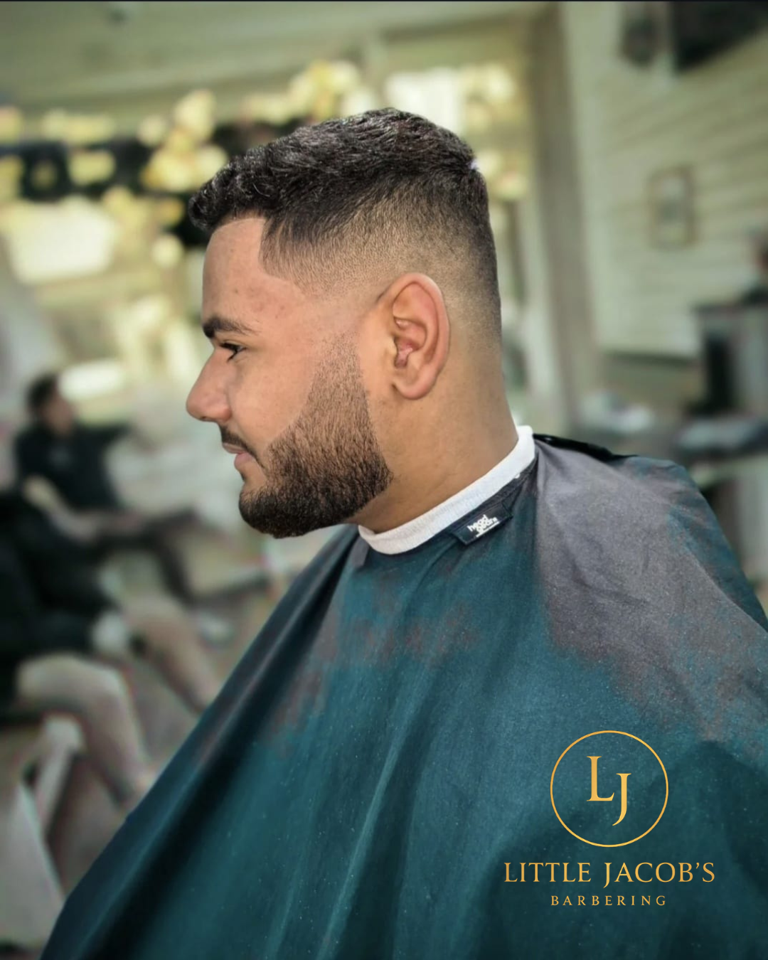 A man with a short, faded haircut and beard sits in a barber's chair at Little Jacob's Barbering. The background shows a barber shop interior.