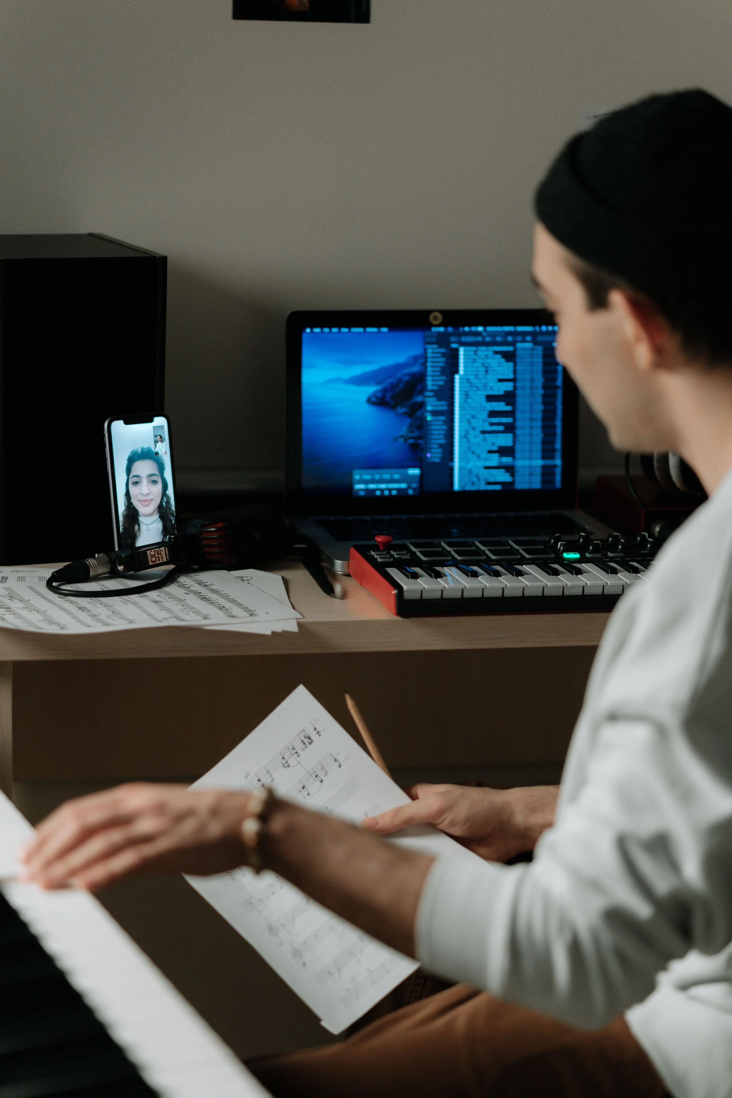 A person wearing a black beanie and a white shirt playing a keyboard, reading sheet music, with a phone showing a woman's face and a computer monitor displaying music production software on their desk.
