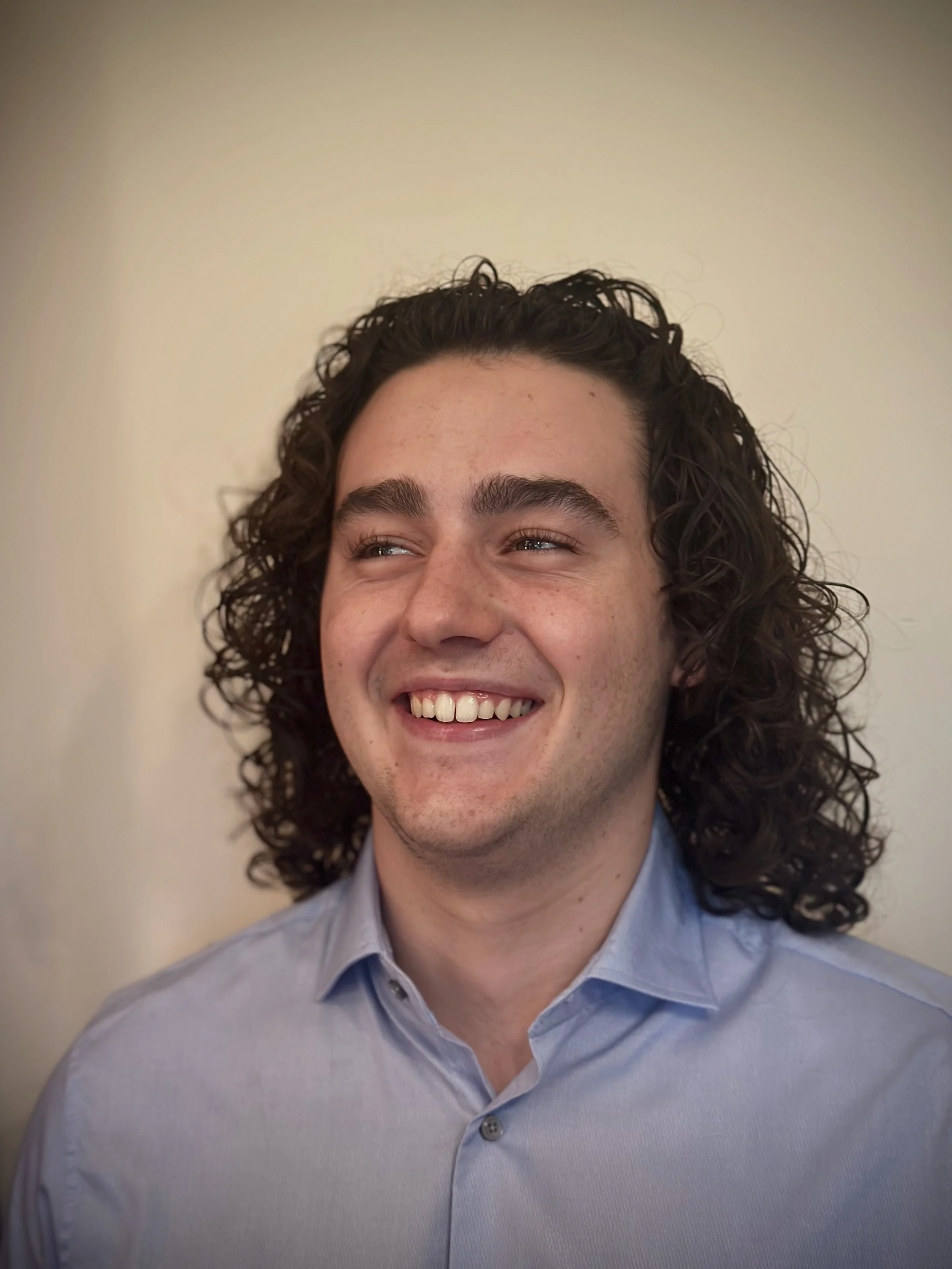 Close-up of a smiling young man with curly dark hair, wearing a light blue collared shirt against a plain light-colored background.