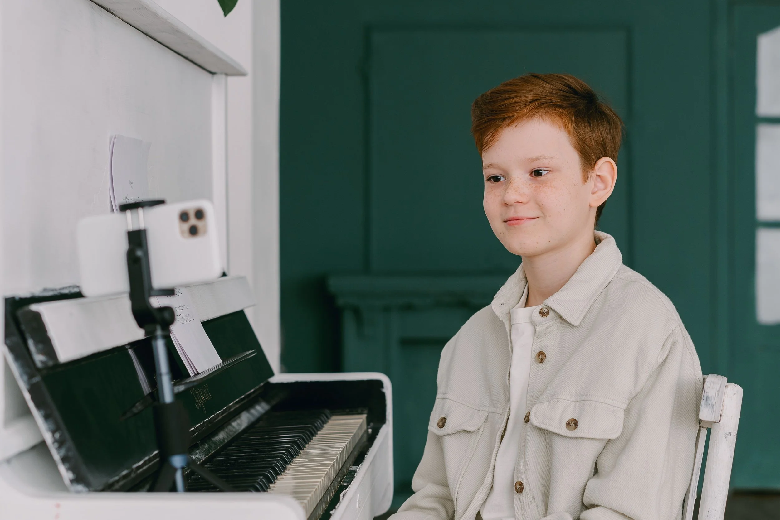 A young boy with brown hair and freckles, wearing a beige jacket over a white shirt, playing a white upright piano.
