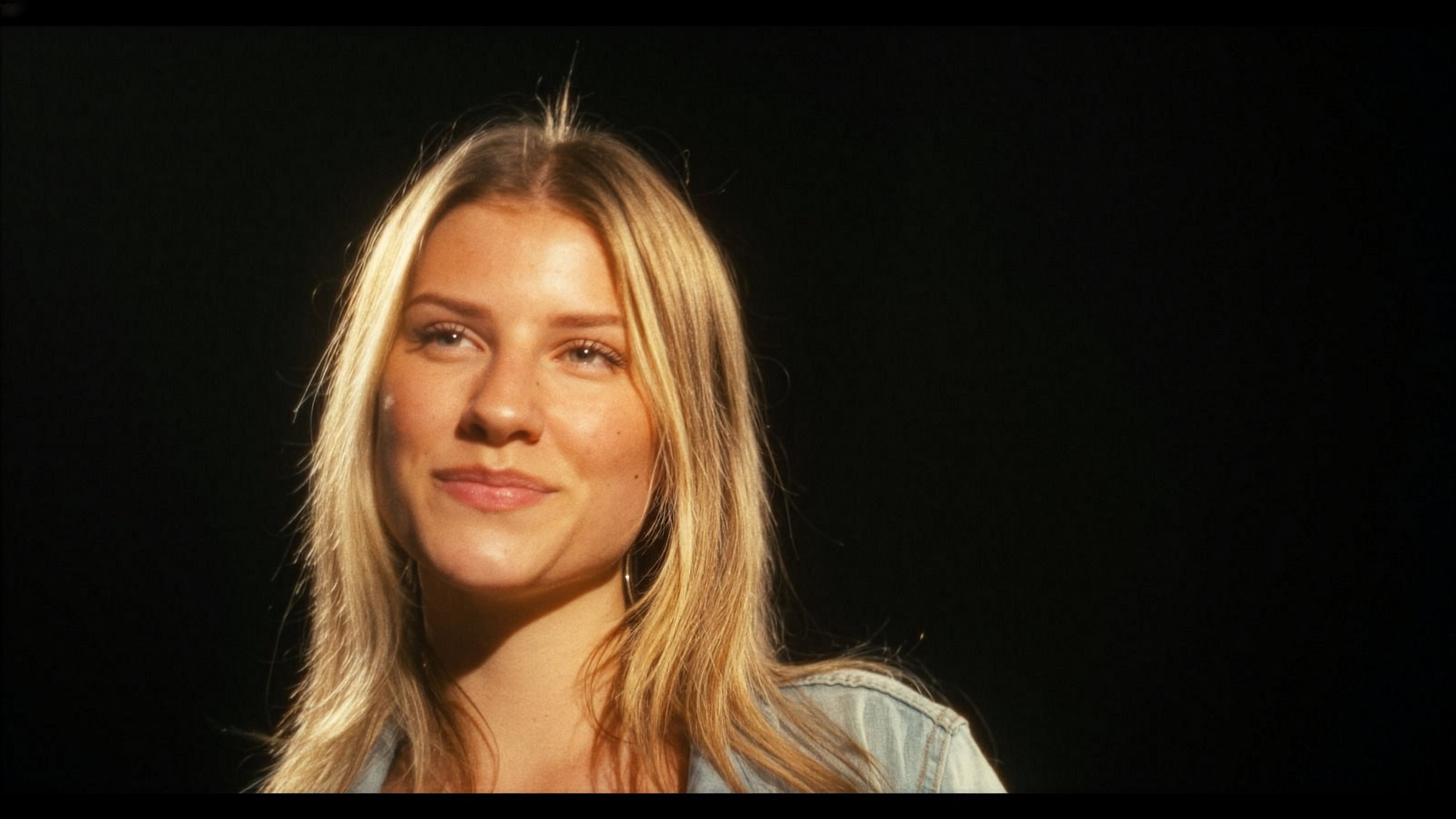 Close-up of a young woman with long, blonde hair and a slight smile standing against a black background.