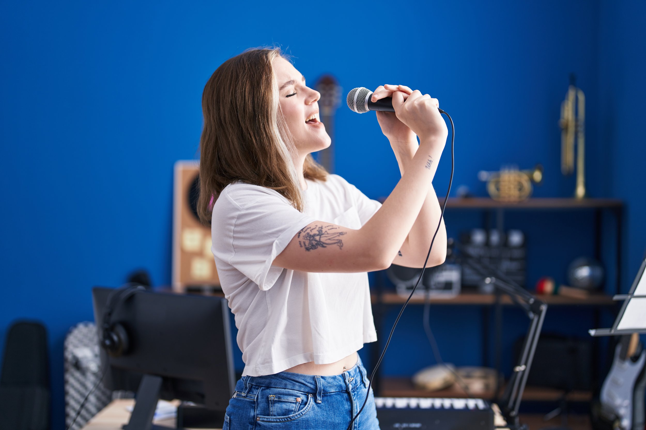 A young woman with long reddish-brown hair singing passionately into a microphone in a room with a bright blue wall.