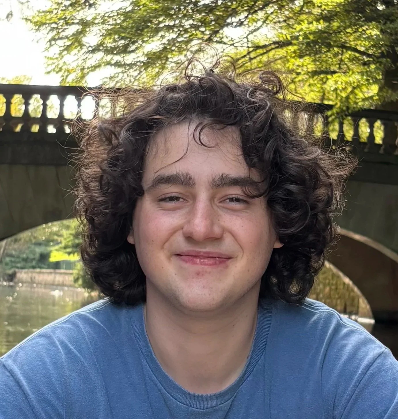 A young man with curly dark hair and a smile, wearing a blue shirt, outdoors near a bridge over a river with green trees in the background.