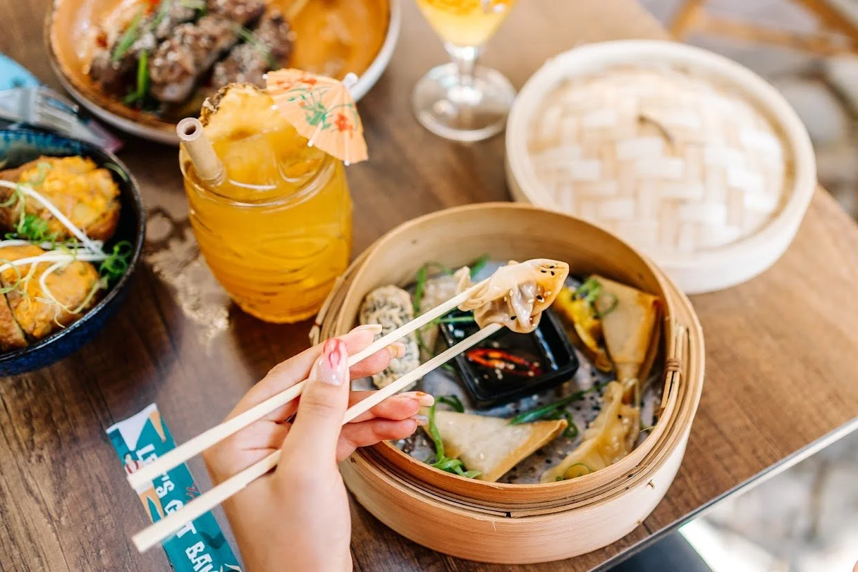 Een person die met eetstokjes een gyoza dumpling vasthoudt boven een ronde Dim Sum stoommandje met andere gyoza's en soja saus. Op de houten tafel staan ook diverse Aziatische gerechten en drankjes.