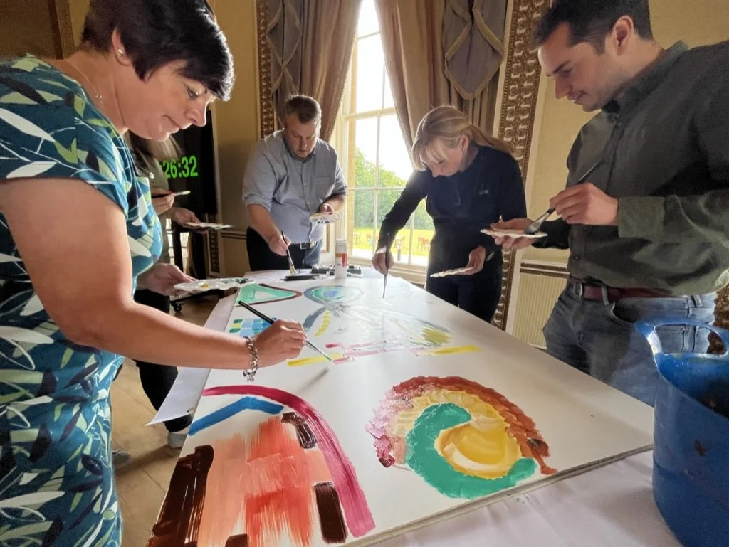 Four people are painting on a large white canvas in a well-lit room with large windows and decorated curtains.