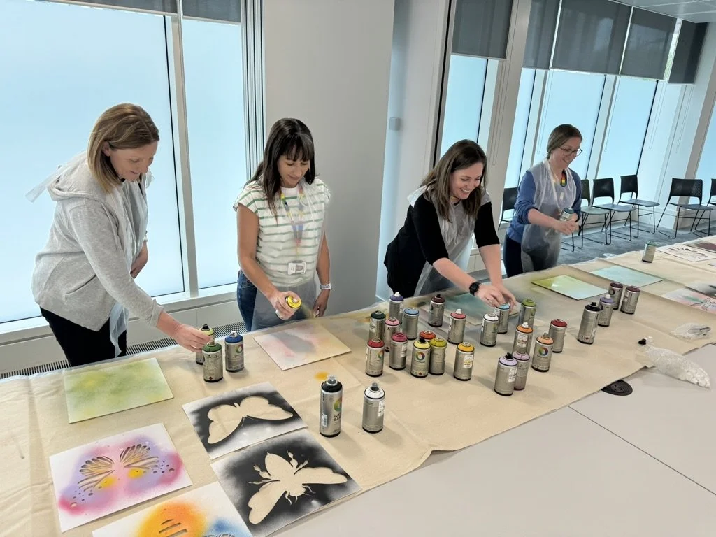 Four women are working on spray painting art projects at a table in a bright room with large windows. There are various spray paint cans and stenciled butterfly and abstract art pieces on the table.