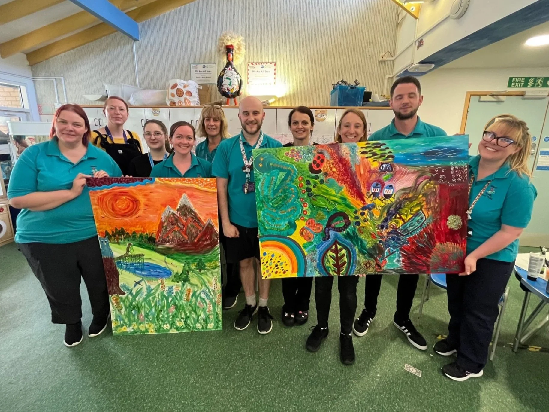 Group of nine people, seven women and two men, standing indoors holding colorful paintings of nature scenes, with a background of shelves, ceiling beams, and a fire exit sign.