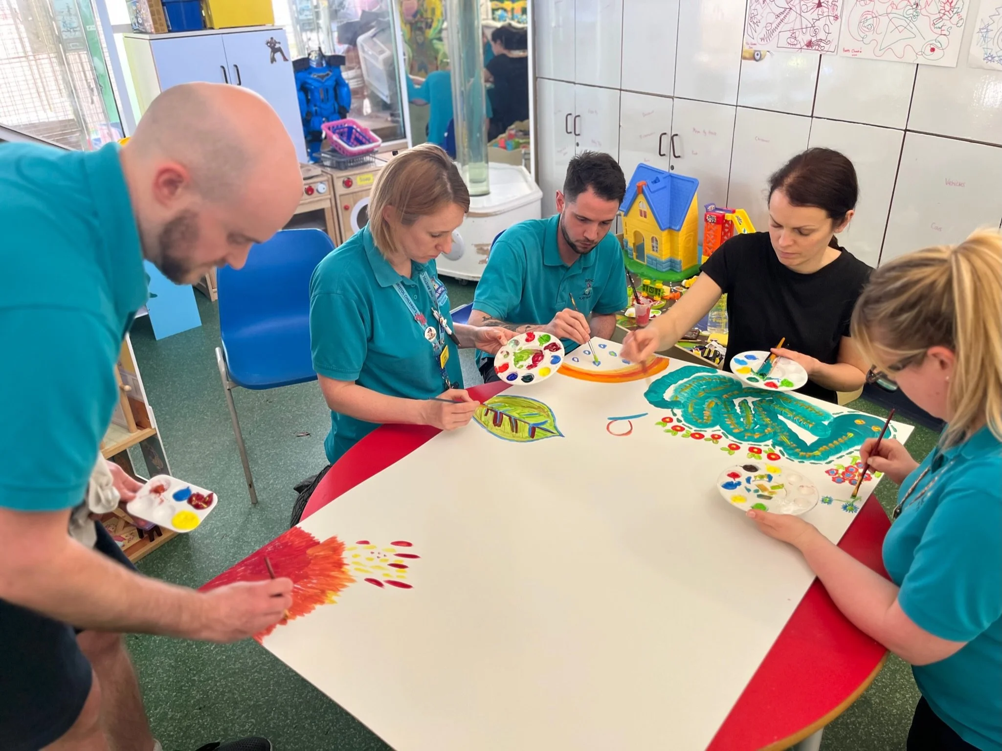 People painting and drawing colorful artwork on large sheets of paper at a table in a classroom.