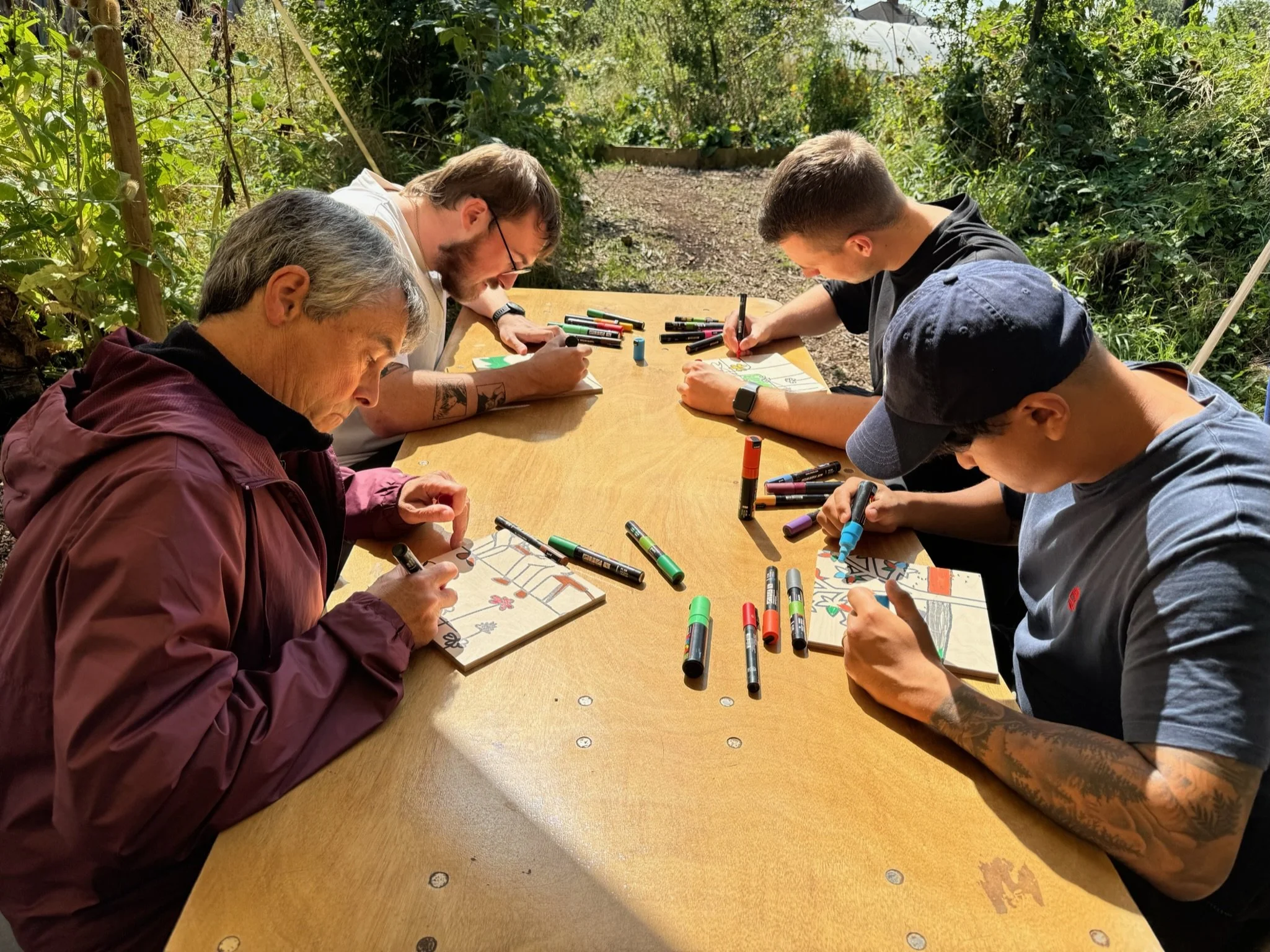 Group of five people drawing and coloring at an outdoor table surrounded by greenery.