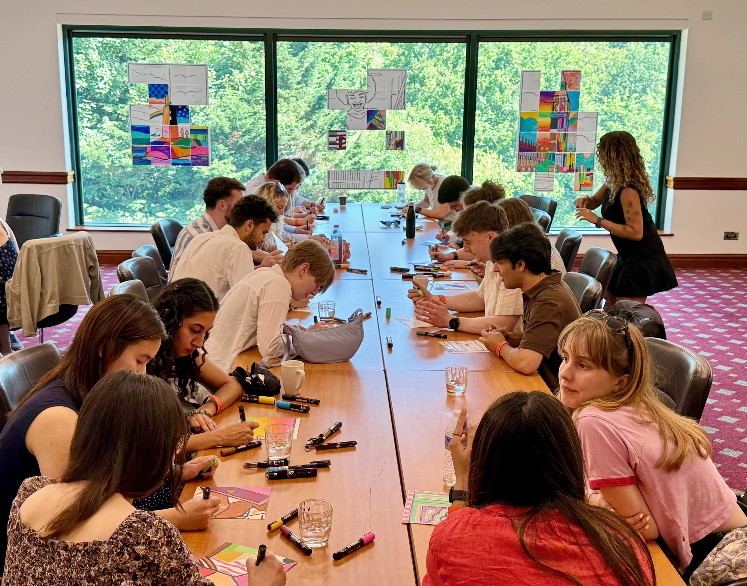 People sitting around a long conference table engaged in coloring activities, with various markers, papers, and drinks on the table, windows showing greenery outside, and illustrations hanging on the window.