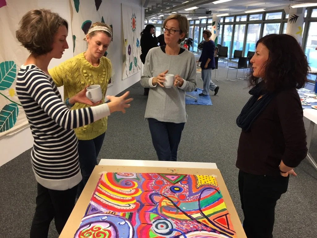 Four women are standing around a table with a colorful abstract painting, engaged in conversation, inside a bright, modern office or gallery space.