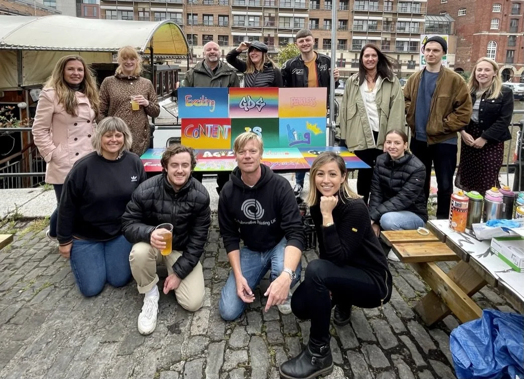 A group of people posing outdoors on a cobblestone area, with a colorful painted signboard behind them and buildings in the background. They are smiling and appear to be celebrating or enjoying a gathering.