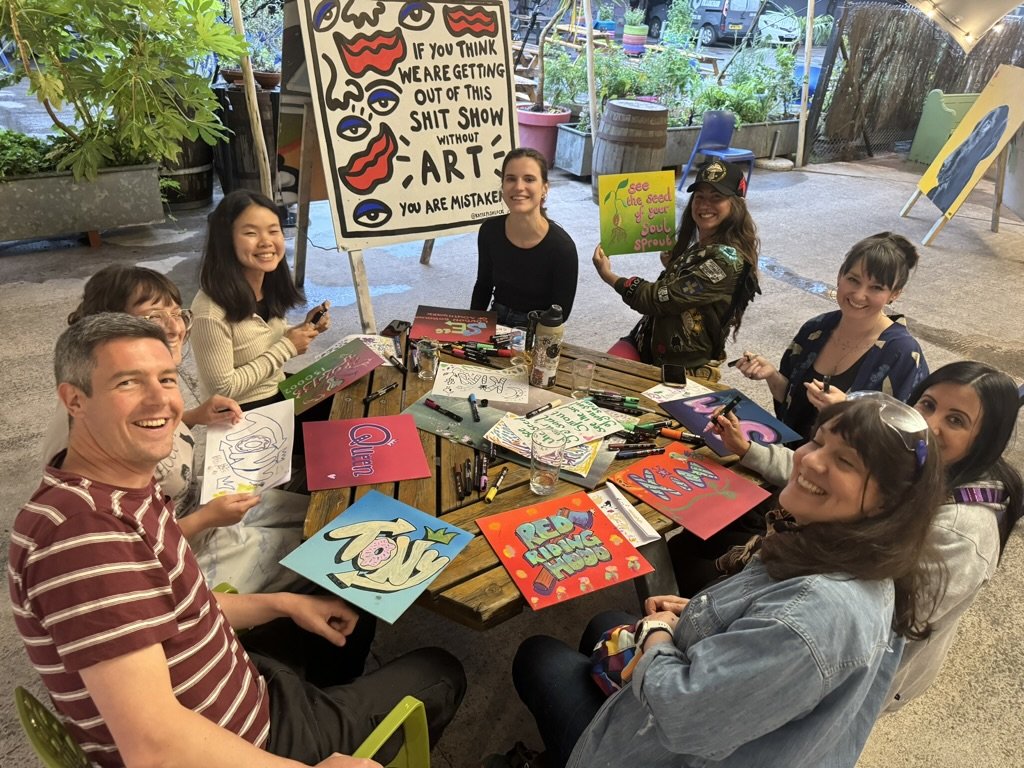 People sitting around a table painting and drawing, with a large art-themed sign in the background, in a bright indoor space with plants and artwork.