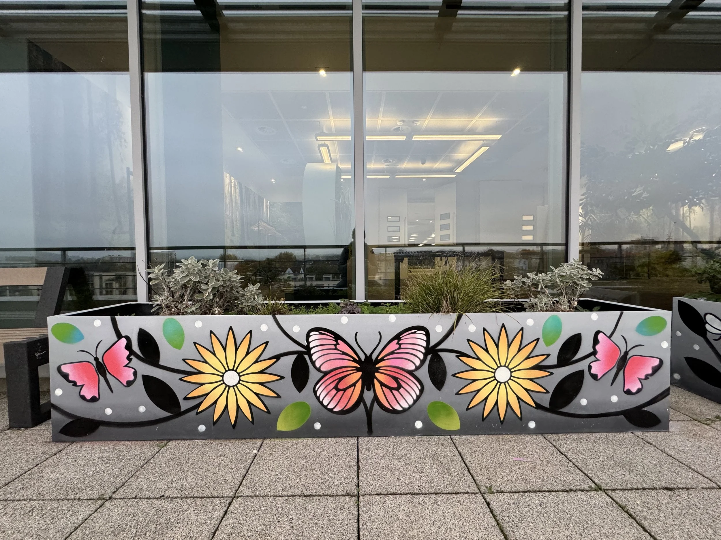 Colorful flower and butterfly artwork on a large planter box in front of a glass building with reflections of the sky and surroundings.