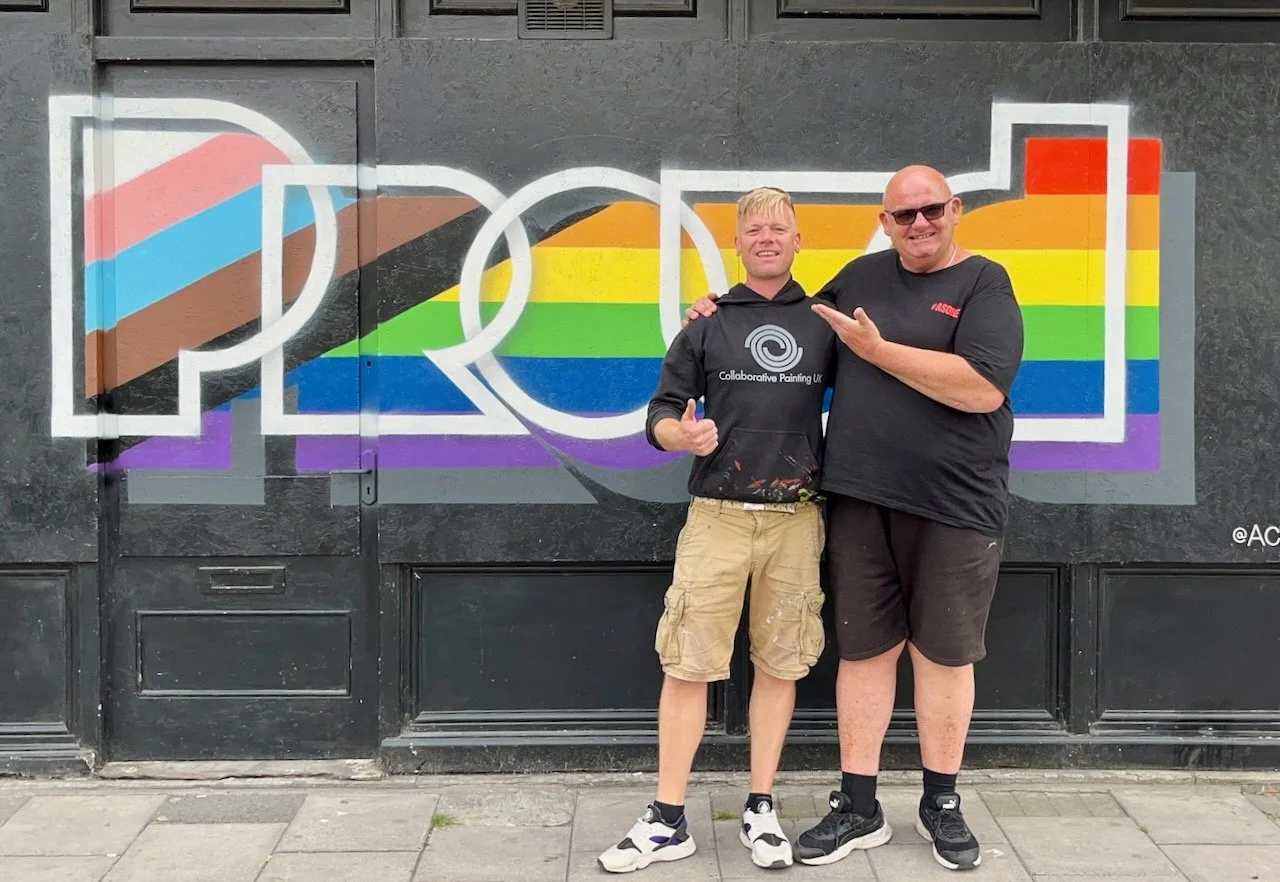 Two men standing in front of a black wall with colorful graffiti spelling 'POOP' in rainbow colors, with one man in a hoodie and the other in a t-shirt, smiling and posing together.