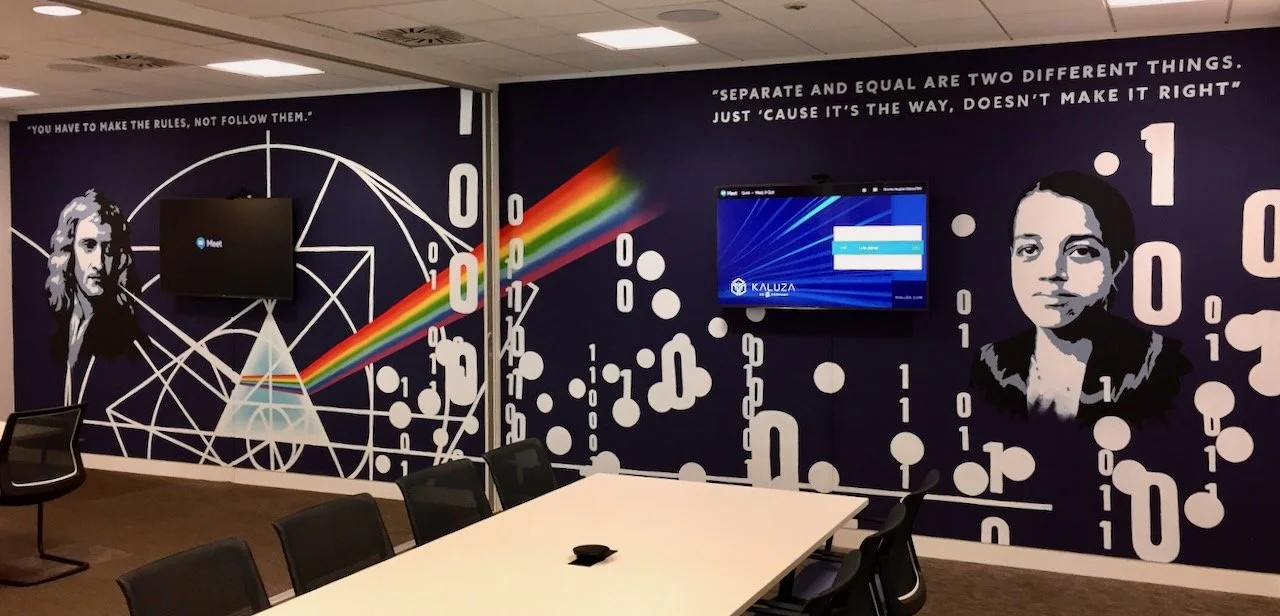 Wall mural with portraits of two women on a navy blue background, surrounded by binary code and geometric shapes, including a rainbow beam. There are two flat-screen monitors mounted on the wall and a conference table with chairs in the foreground.