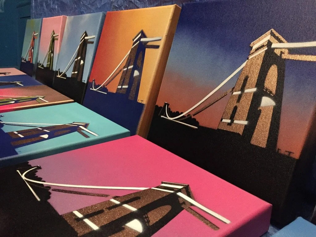 Multiple colorful canvases of bridge paintings with different sunset backgrounds, arranged on a table.