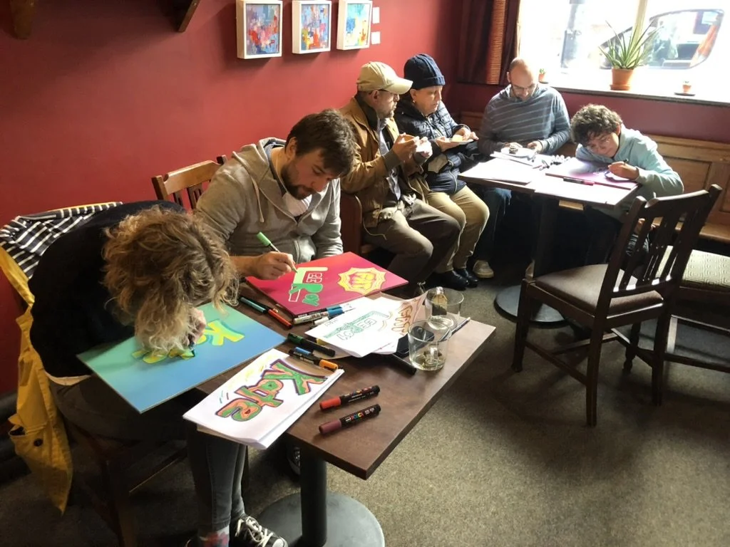Group of people, including children and adults, sitting at tables in a cozy cafe or art studio, engaging in drawing and coloring artwork.