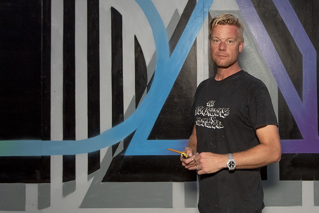 A man with light hair and a beard, wearing a black T-shirt and silver watch, stands in front of a colorful geometric mural. He holds a small orange paintbrush in his right hand.