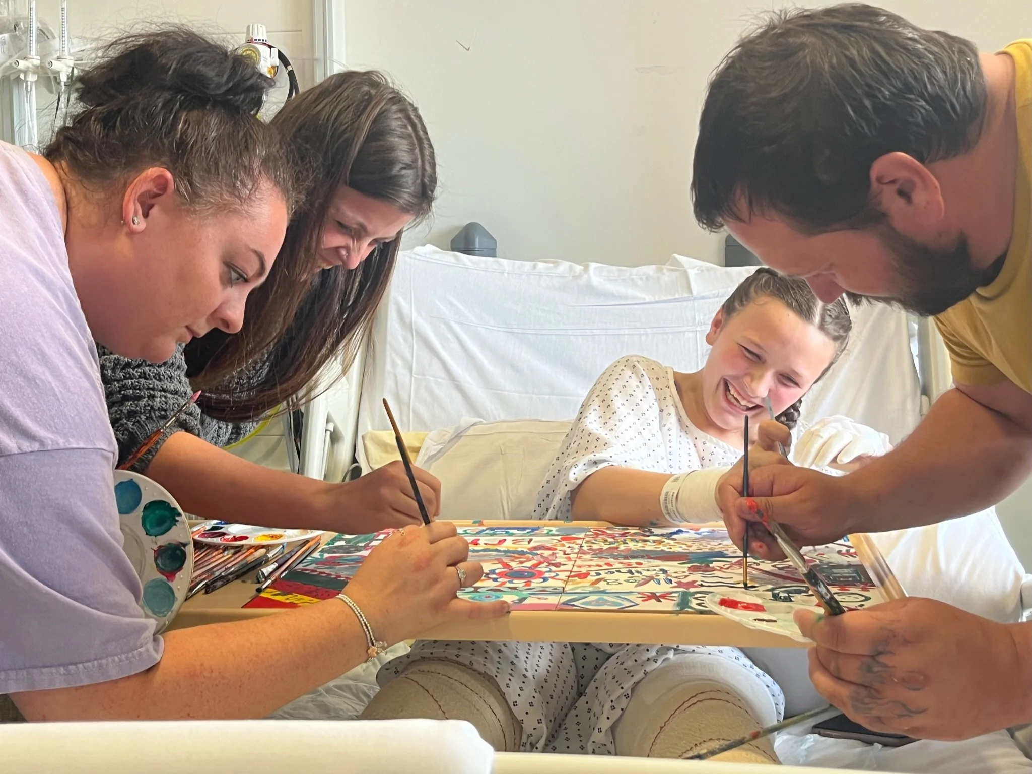 A group of four people, including three adults and one young girl, are gathered around a table in a hospital room, painting on a large colorful poster or canvas with brushes and paint palettes, smiling and engaging in an art activity.
