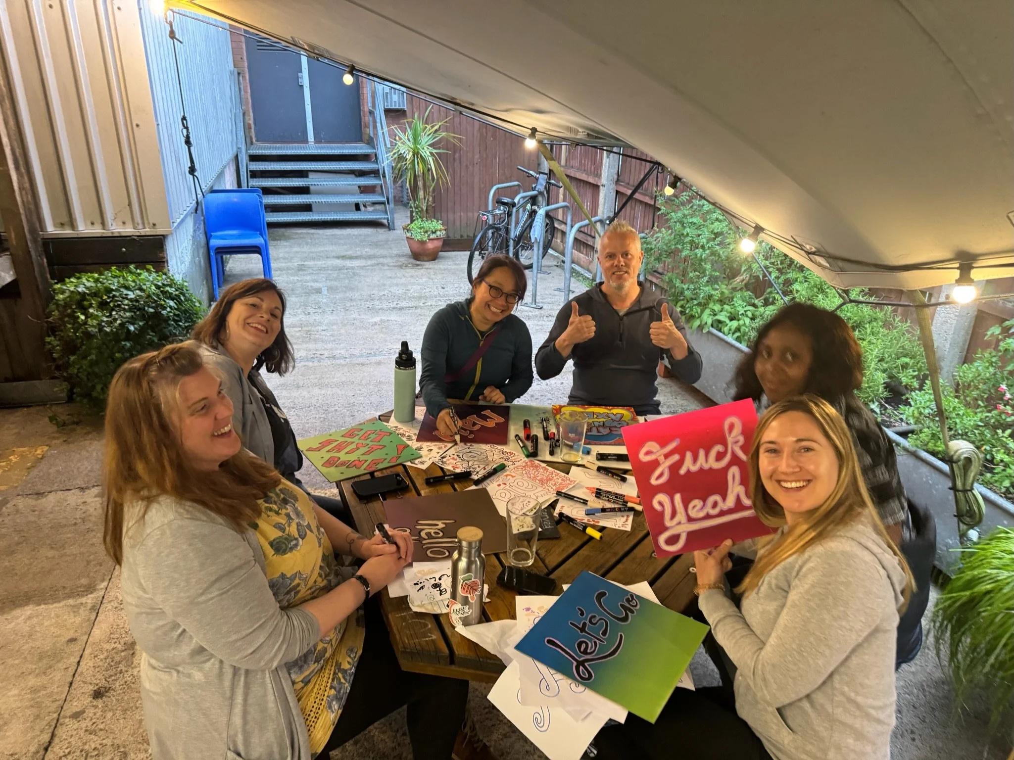 A group of people sitting at a table outdoors under a canopy, engaging in arts and crafts activities with colorful signs and markers, smiling and enjoying the event.