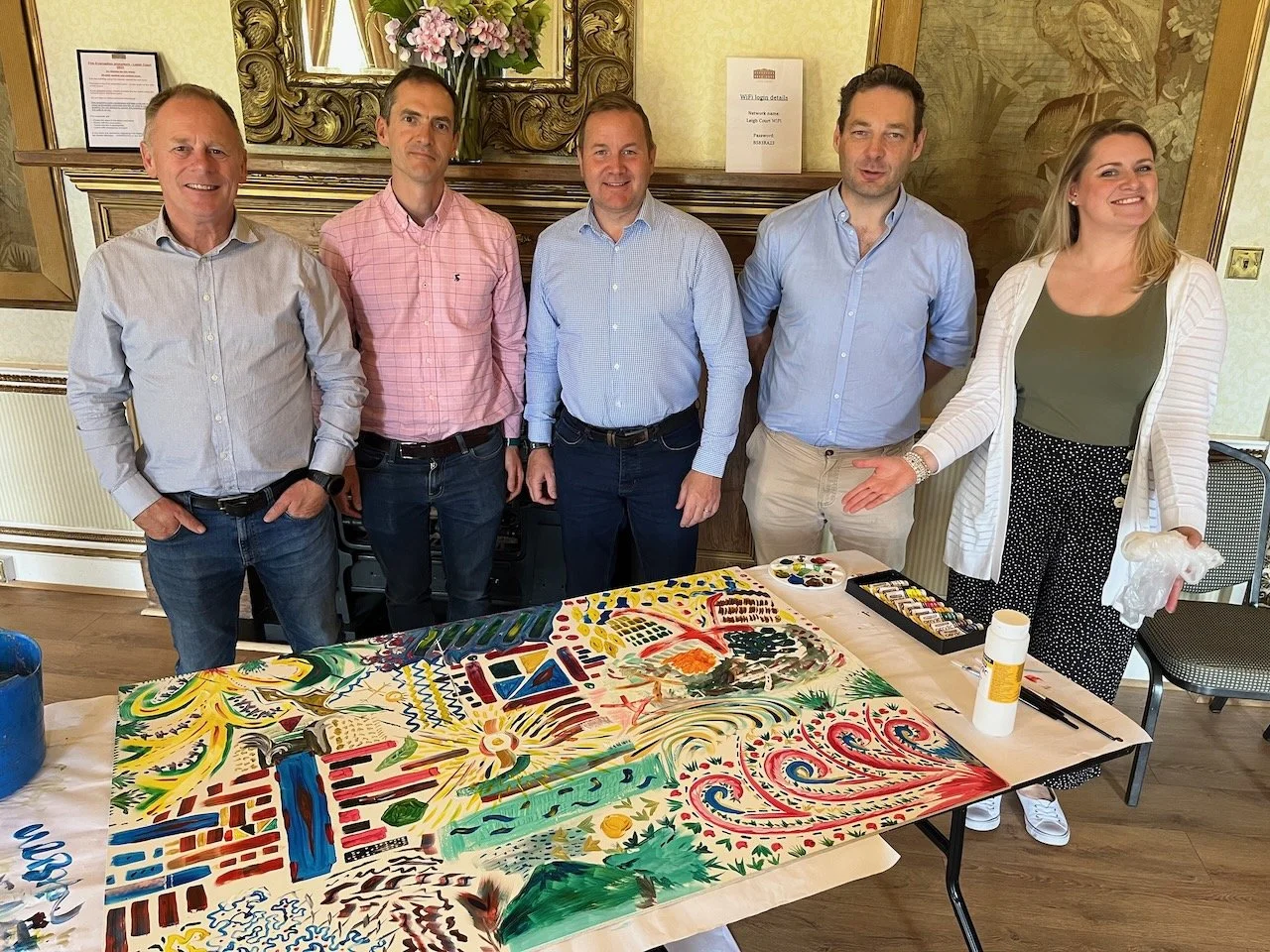 Five people, four men and one woman, standing behind a large colorful abstract painting on a table, in a room with ornate decor and a mirror with flowers above it.