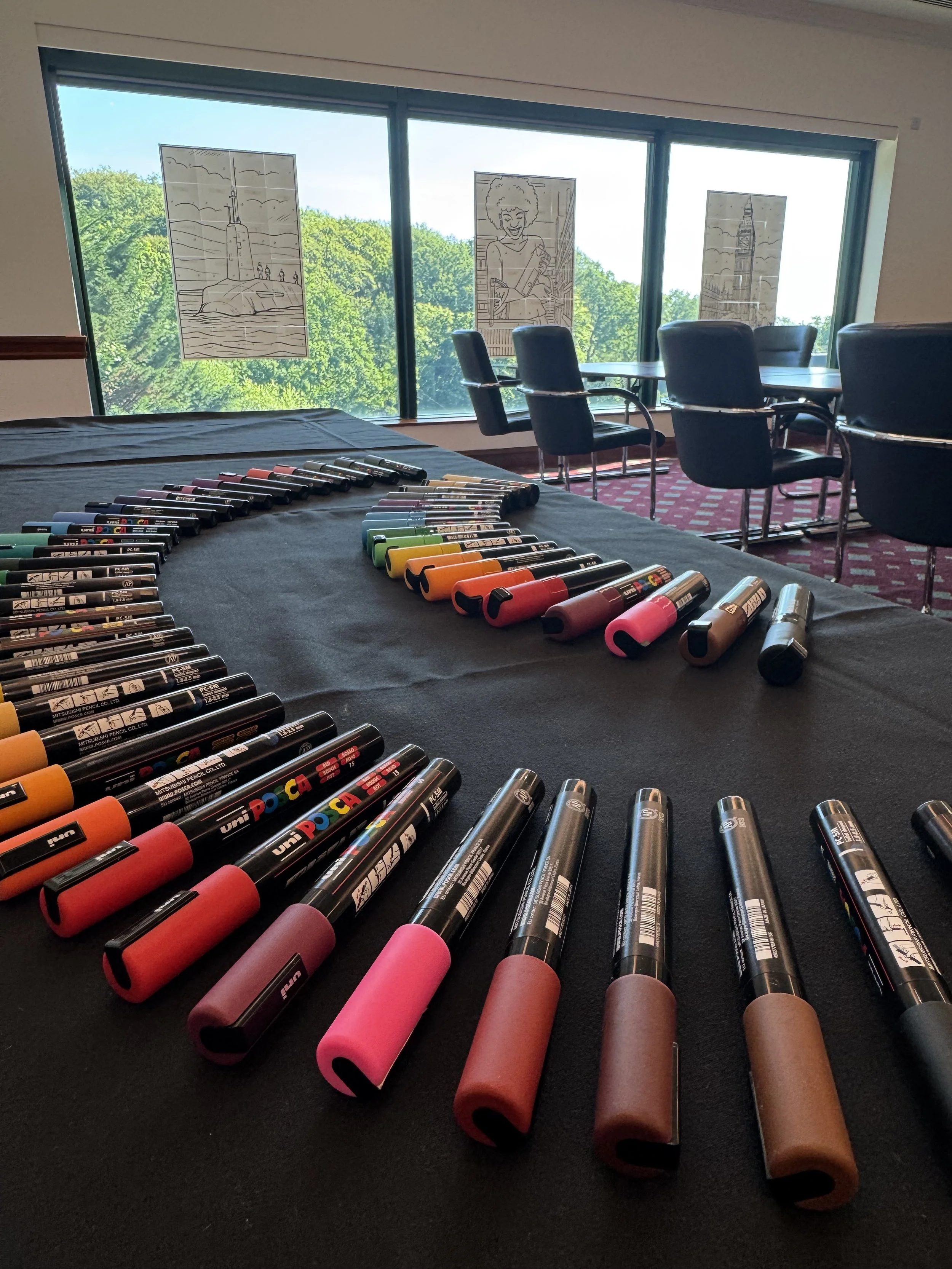 Colorful markers arranged on a black table in a conference room with table and chairs, with windows showing green trees outside, and black-and-white line art posters on the window glass.