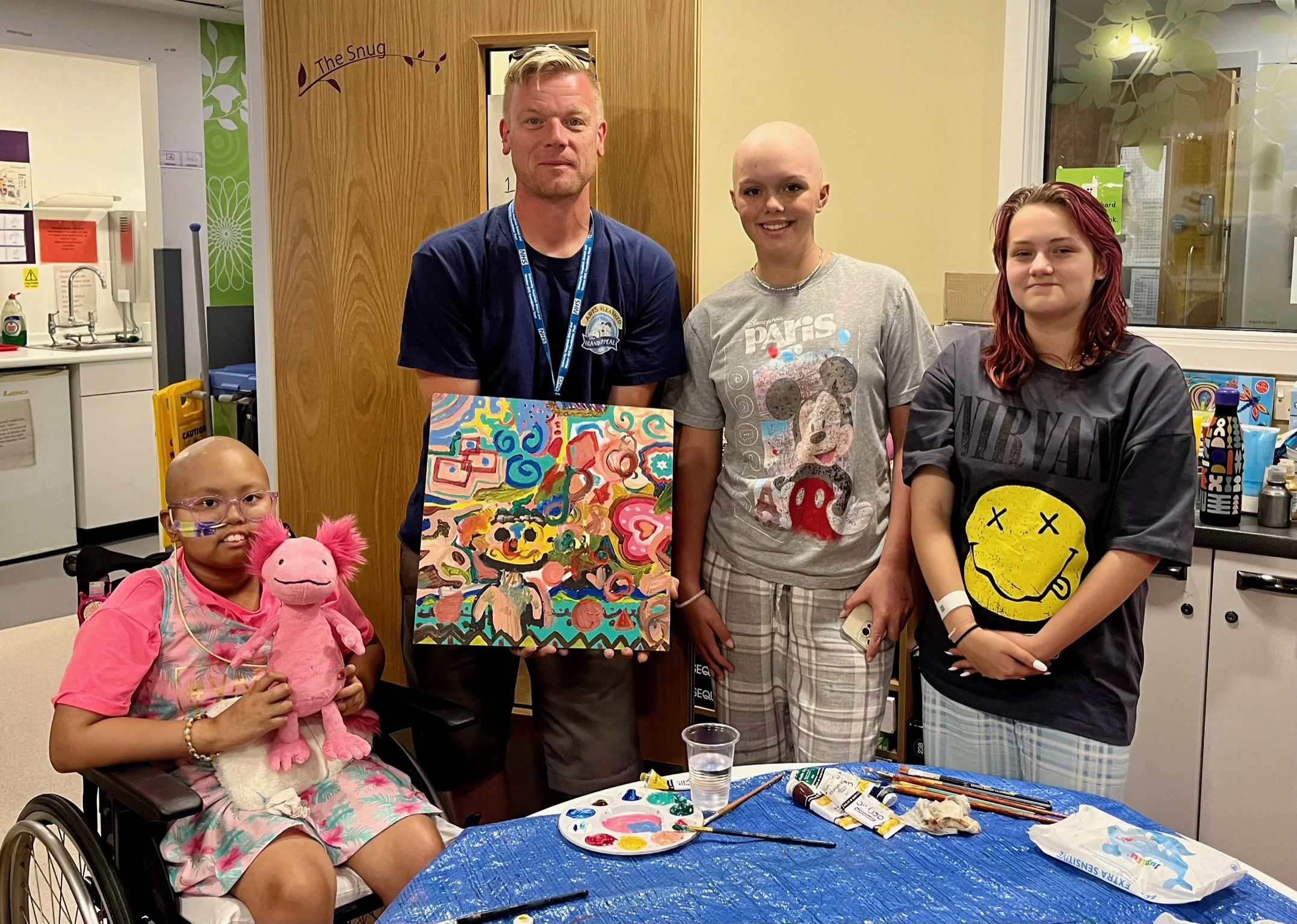Group of five people, including a girl in a wheelchair holding a pink plush pig, gathered around an art table with paint supplies. Two women and two men stand behind her, one of whom is presenting a colorful abstract painting. The setting appears to 